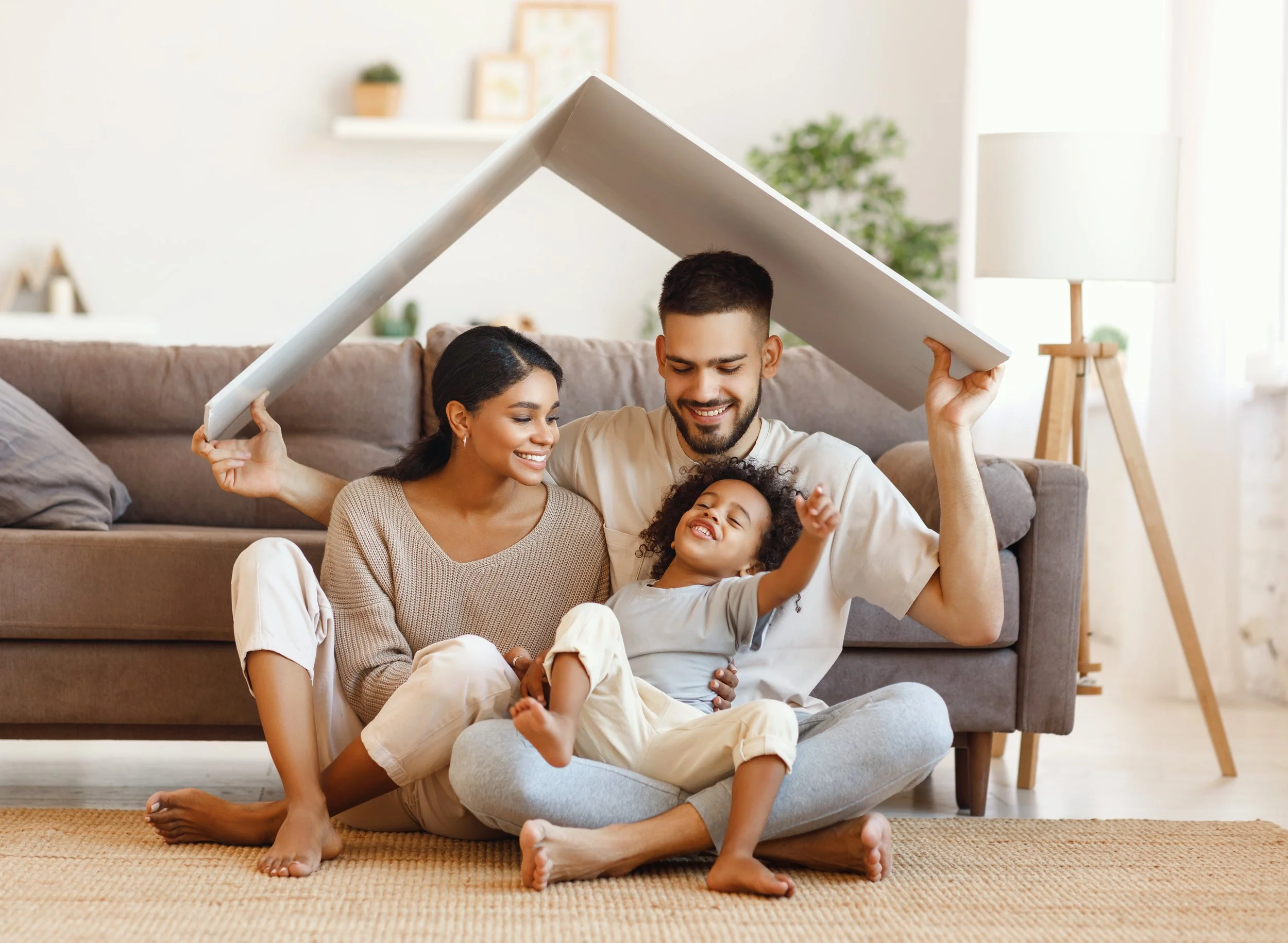 A family of four sitting on a carpeted floor inside a living room, with a white table and beige sofa behind them, playing a game with a large white foam board kept over their heads, smiling and laughing together.