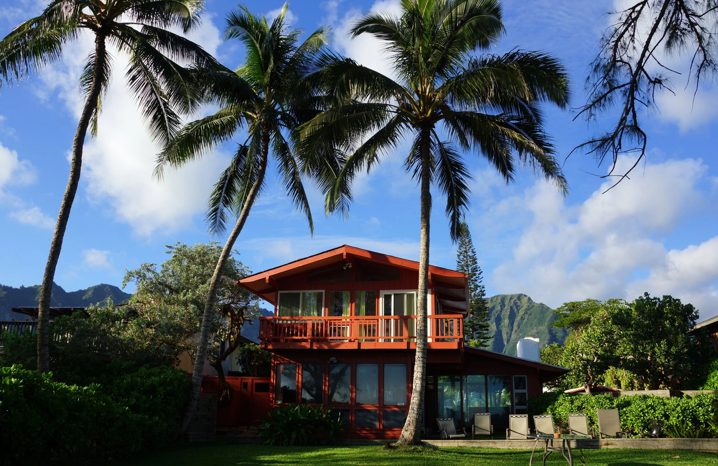 A multi-level vacation house painted in red with large glass windows and a balcony, surrounded by tall palm trees, green bushes, and chairs outdoors, with mountains in the background under a partly cloudy blue sky.