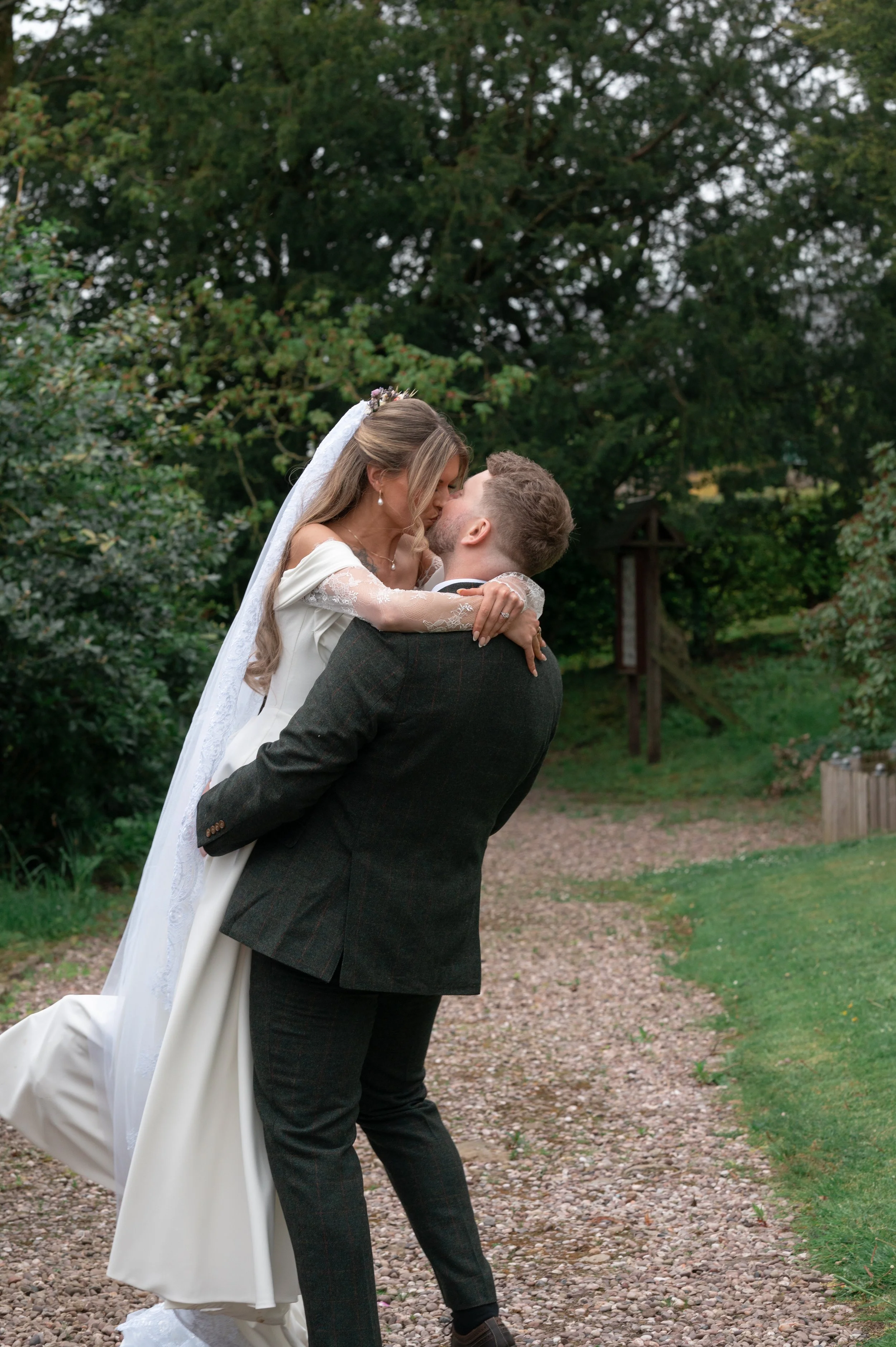 A bride and groom sharing a kiss outdoors in a garden, with the groom lifting the bride. The bride is wearing a white wedding gown with lace sleeves and a veil, while the groom is dressed in a dark suit. Green trees and bushes are in the background.