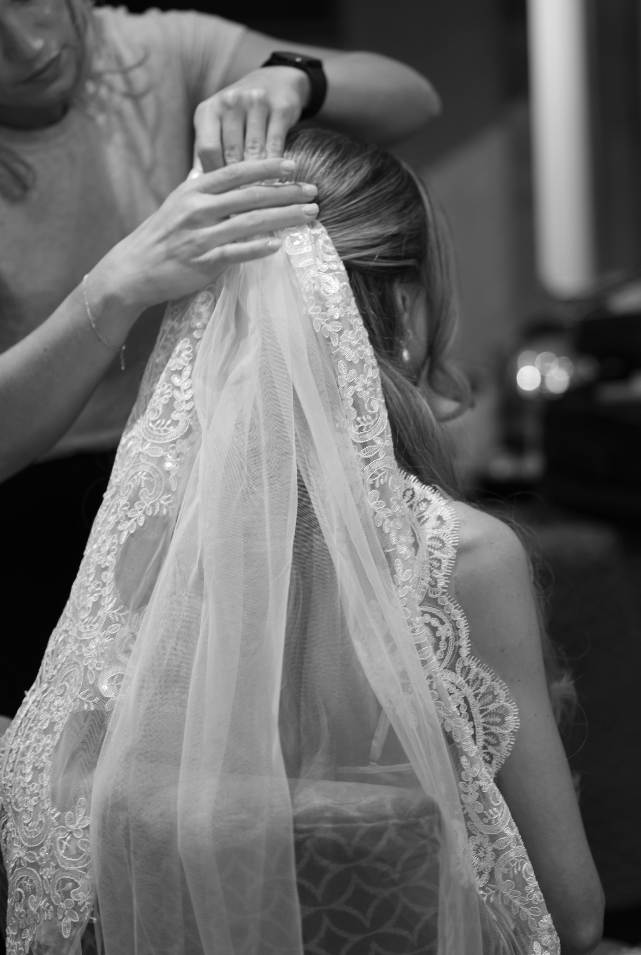 A person helping a bride with her wedding veil, showing close-up of hands adjusting the lace veil over the bride's head.