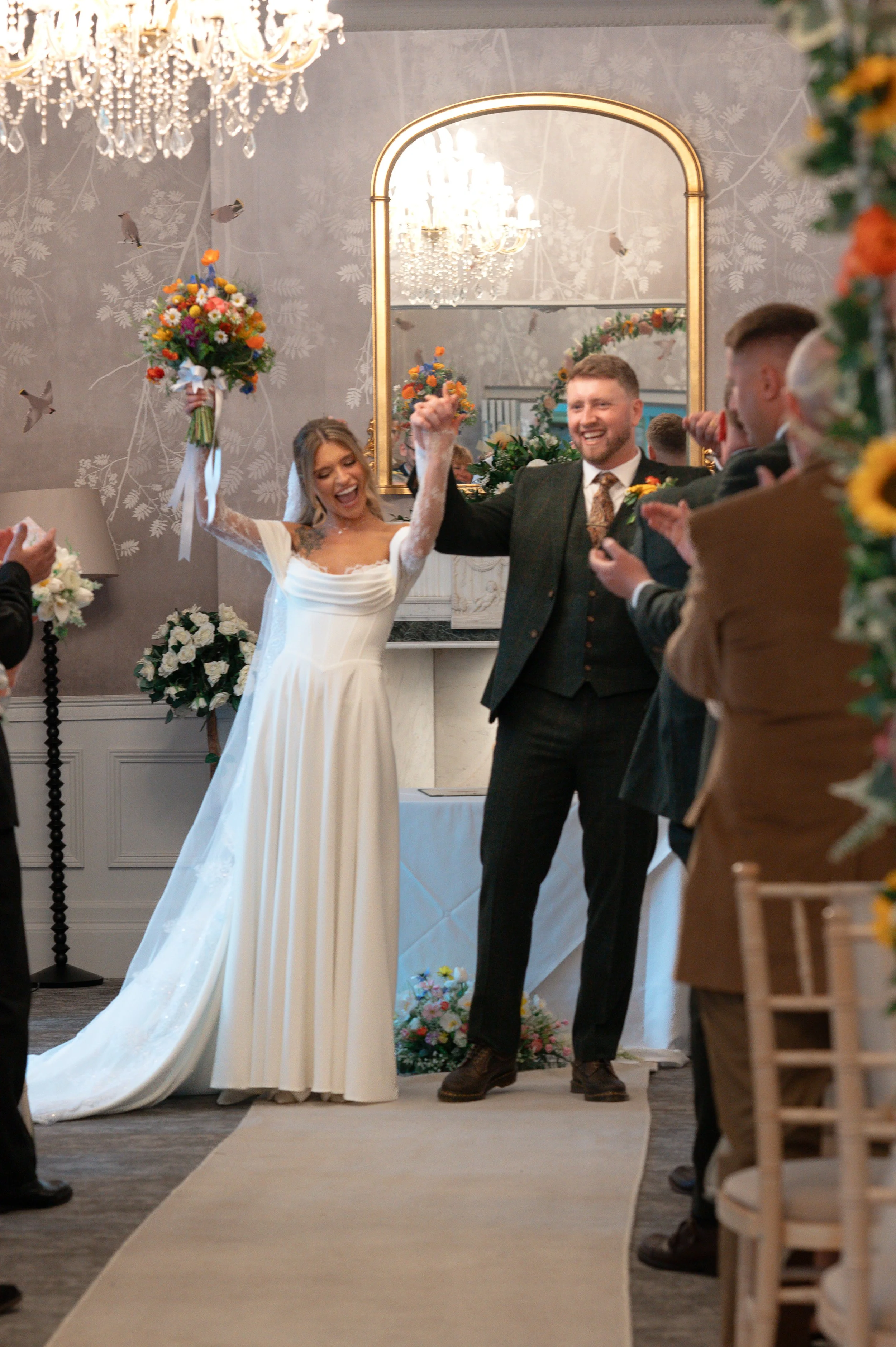 A bride and groom standing together in a decorated wedding venue, surrounded by friends and family, with bouquets of flowers, a chandelier, and a large mirror in the background.