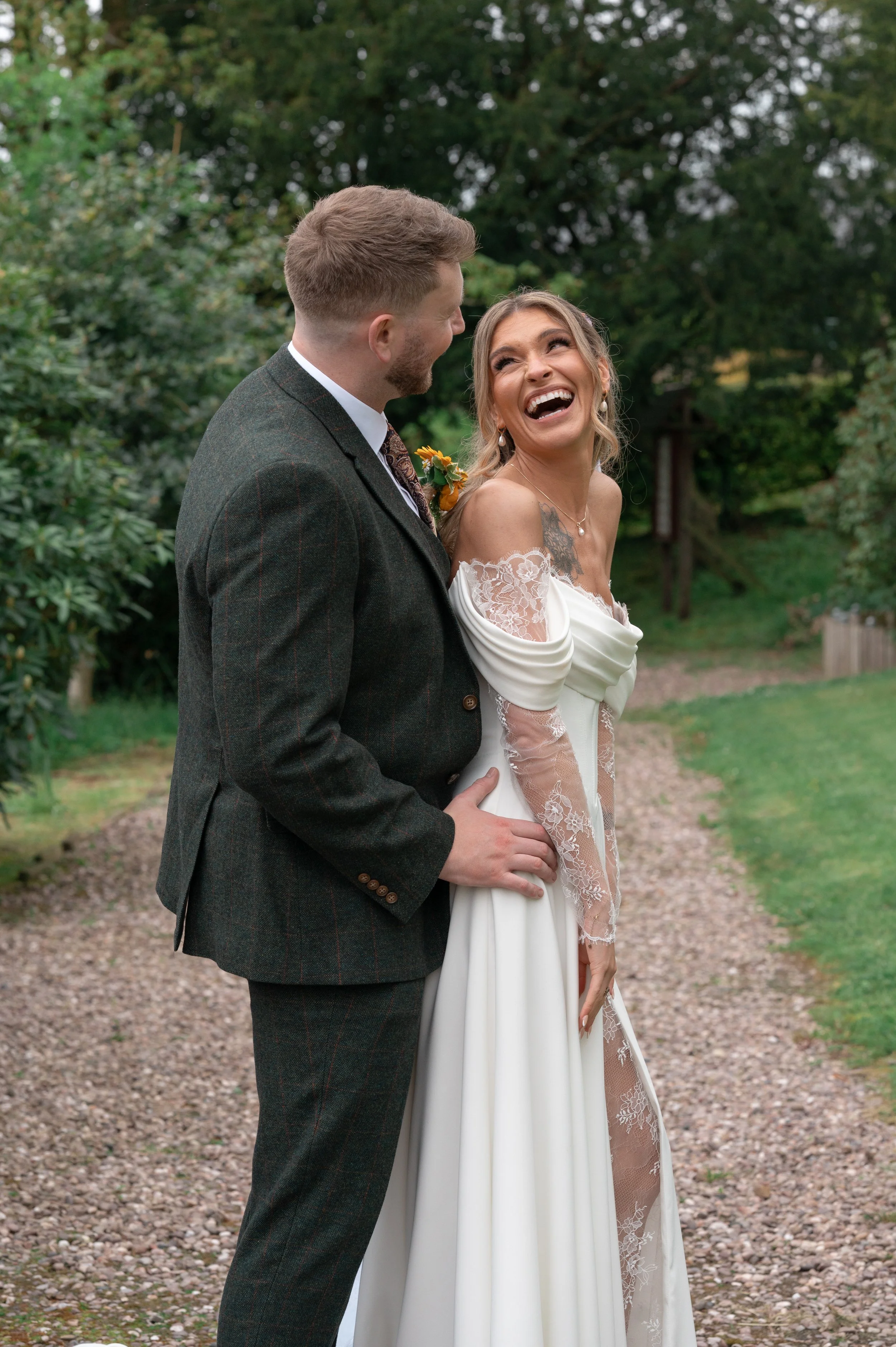 A man and a woman, likely newlyweds, are standing outdoors in a garden with green trees in the background. The woman is wearing a white off-shoulder wedding dress with lace details and the man is in a dark plaid suit. They are laughing and looking at