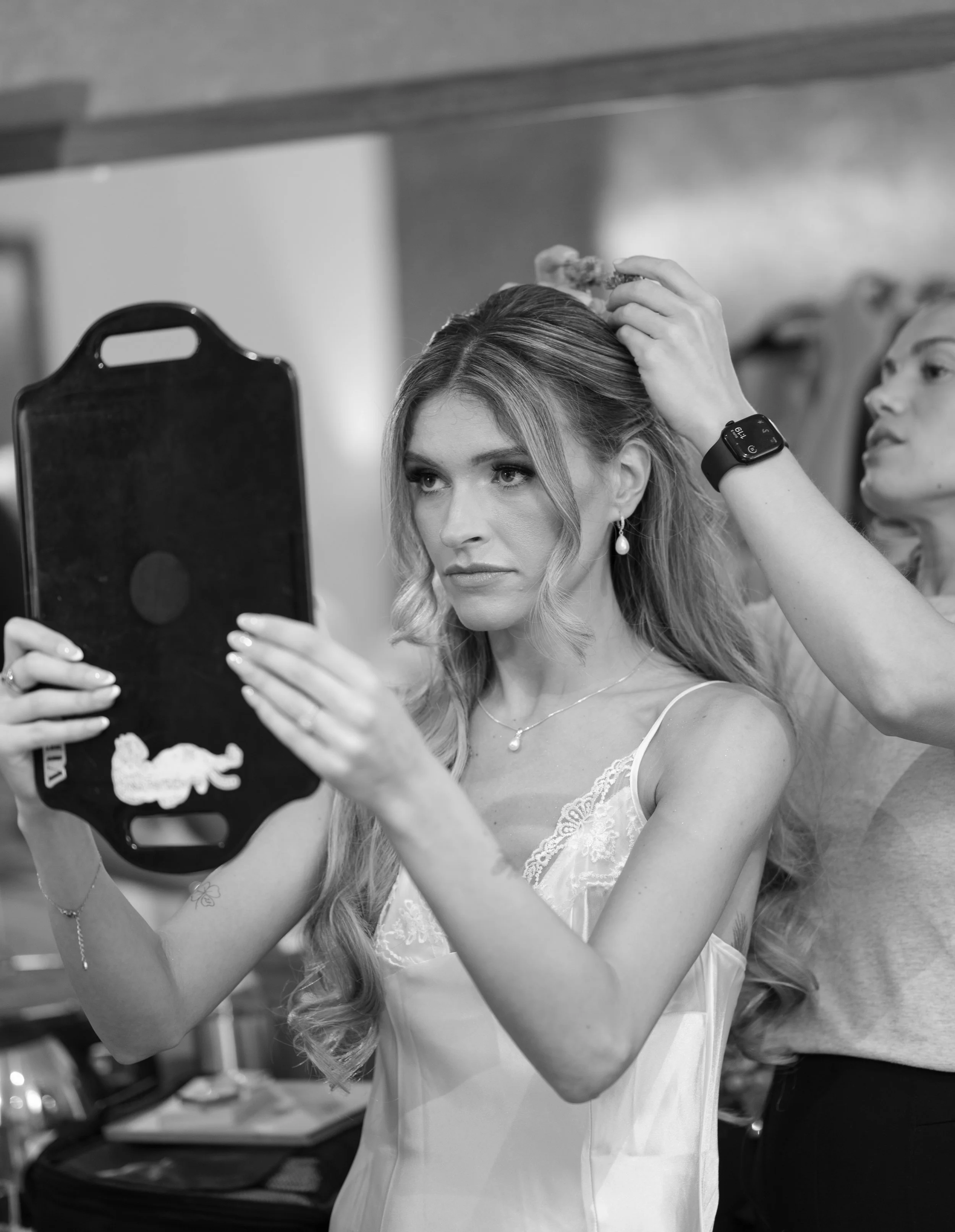 A bride with long, wavy hair is getting her hair styled by a hairstylist at a preparatory venue. The bride is wearing earrings, a delicate necklace, and a white satin or silk dress with lace details.
