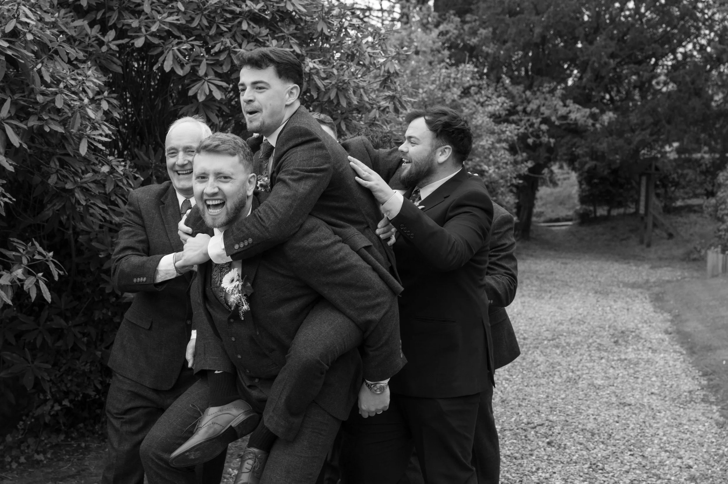 Group of men wearing suits having fun outdoors, one man is riding on the backs of others, all are smiling and laughing in a park-like setting.