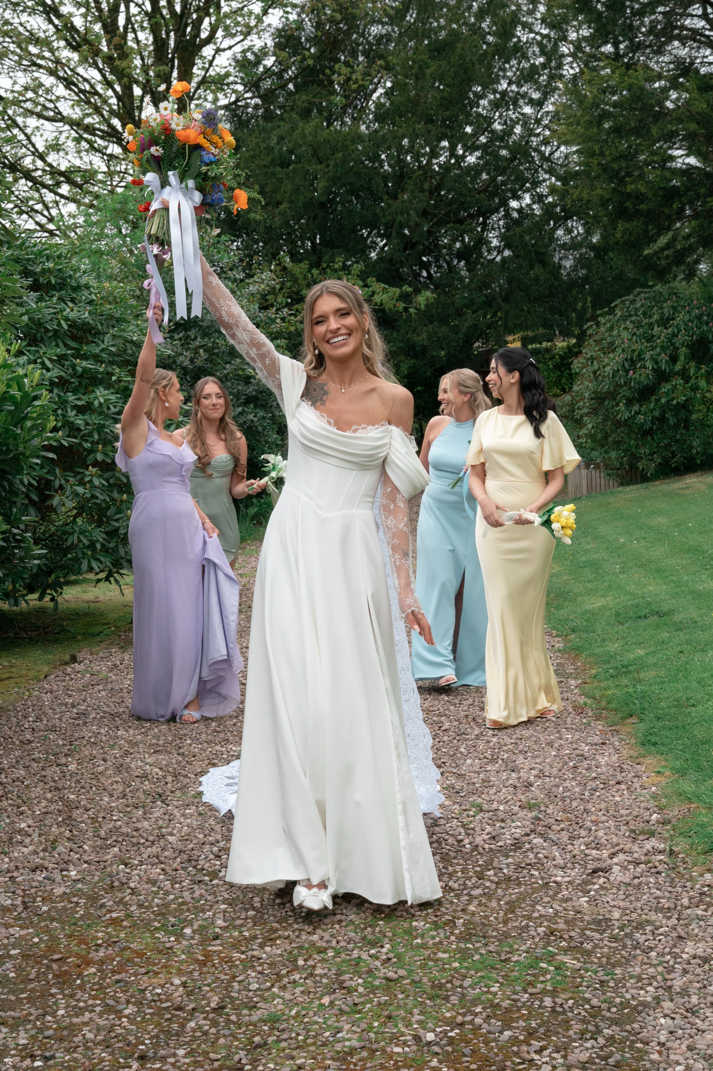 A bride in a white wedding dress holding a bouquet of flowers above her head, surrounded by bridesmaids in pastel-colored dresses outdoors on a gravel path with trees in the background.