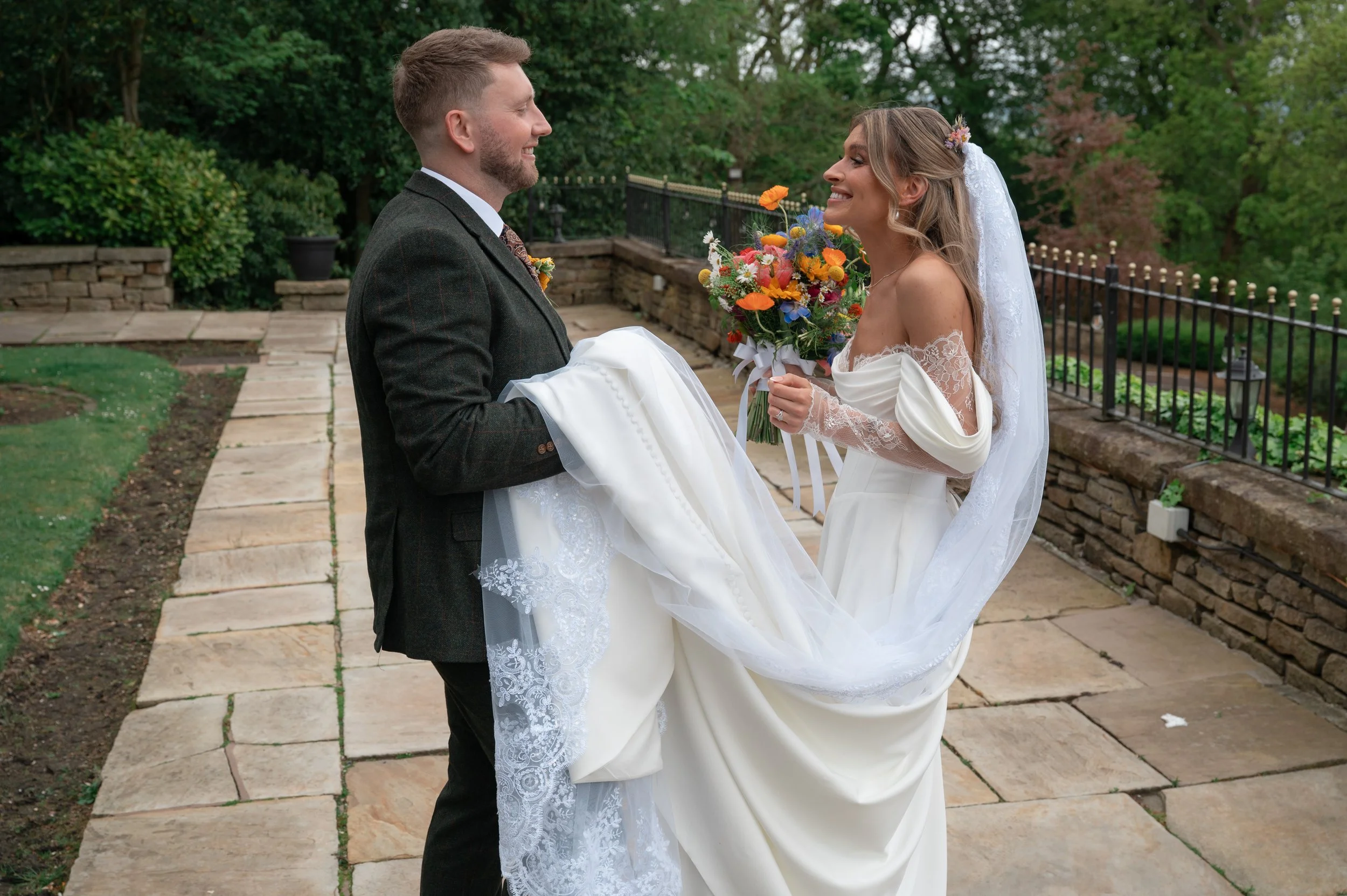 A bride and groom smiling at each other outdoors, with the groom carrying the bride's wedding dress. The bride holds a colorful bouquet and wears a lace wedding gown and veil. The background shows a stone patio, green trees, and a decorative metal fe