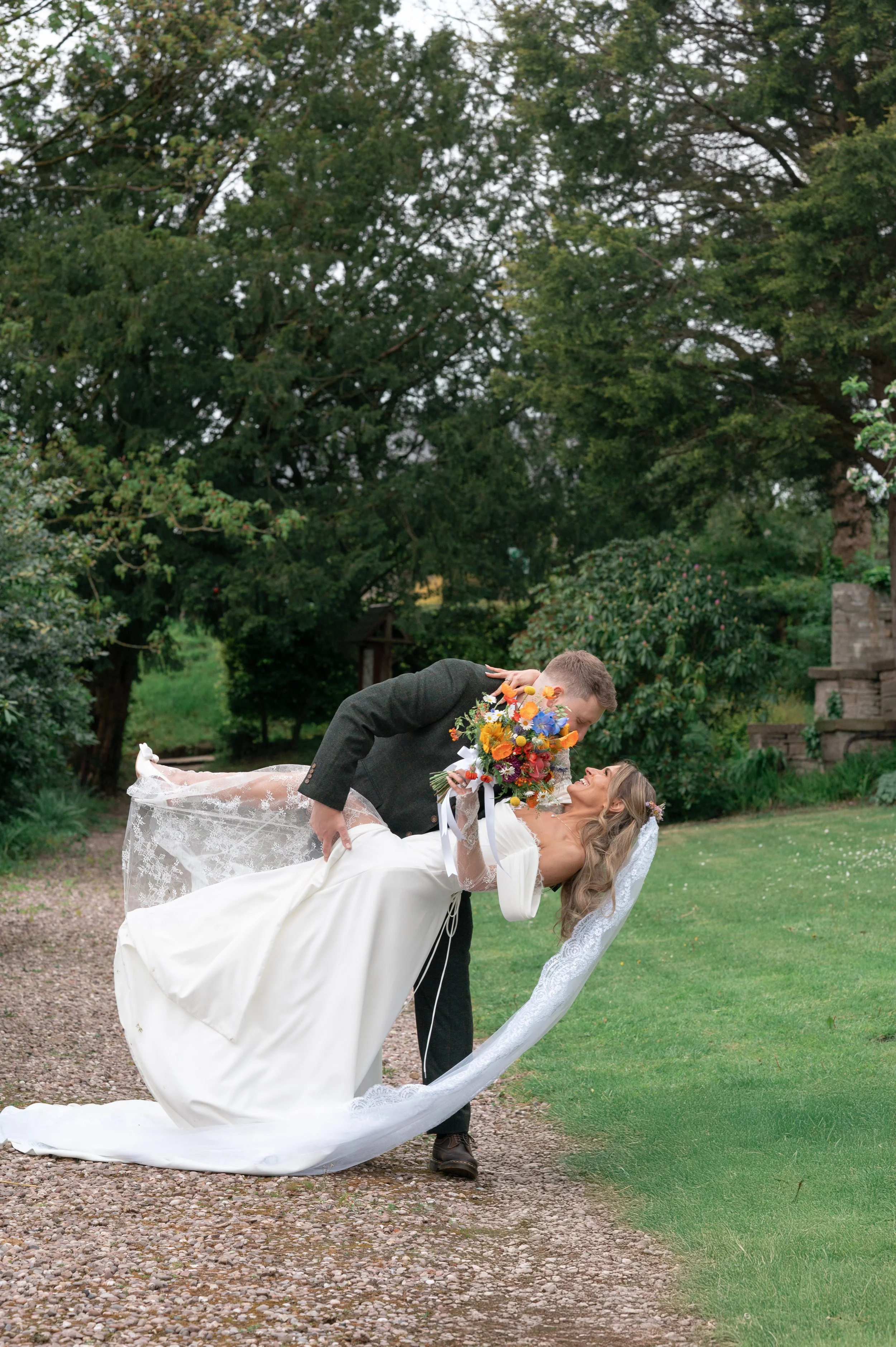 A bride is holding a bouquet of colorful flowers while a groom leans over her, both smiling, outdoors in a garden setting with trees and greenery.