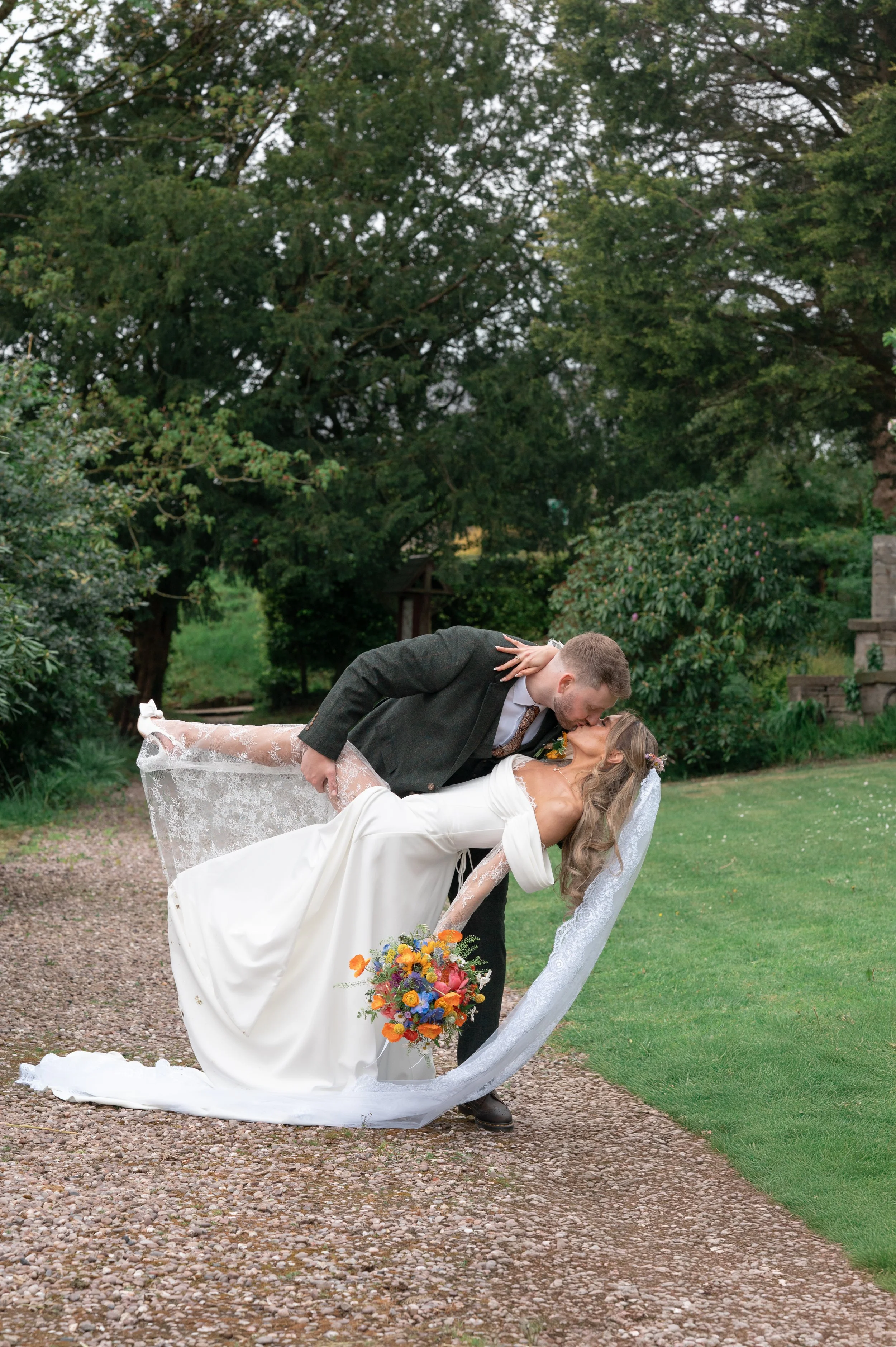 A newlywed couple sharing a kiss outdoors. The groom is dipping the bride, who holds a colorful bouquet, during daytime with green trees in the background.