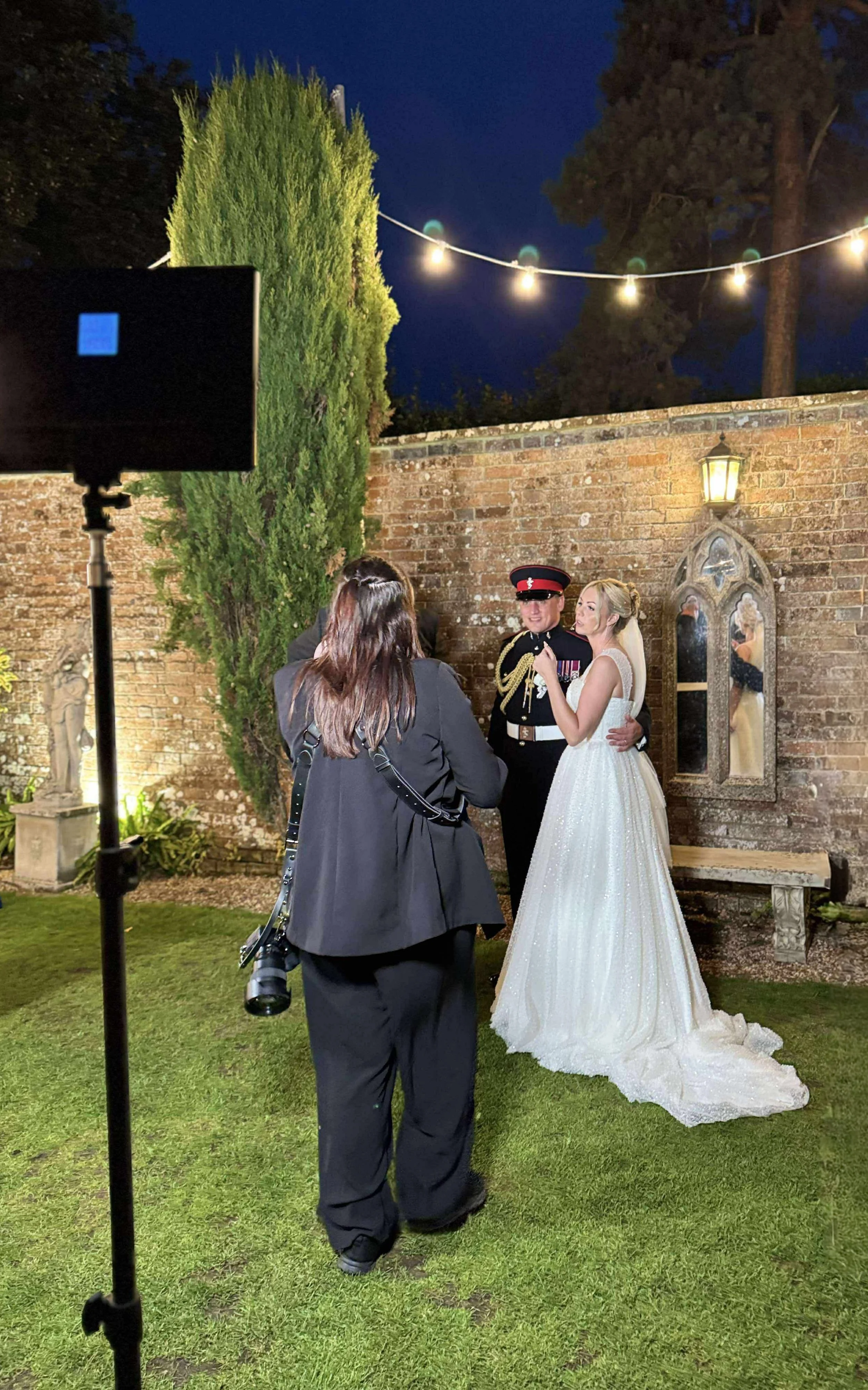 A bride in a white wedding dress with a long train stands next to a groom in a military uniform during a wedding ceremony at night. They are in an outdoor garden with a brick wall and stained glass window behind them, illuminated by a nearby lantern and string lights.