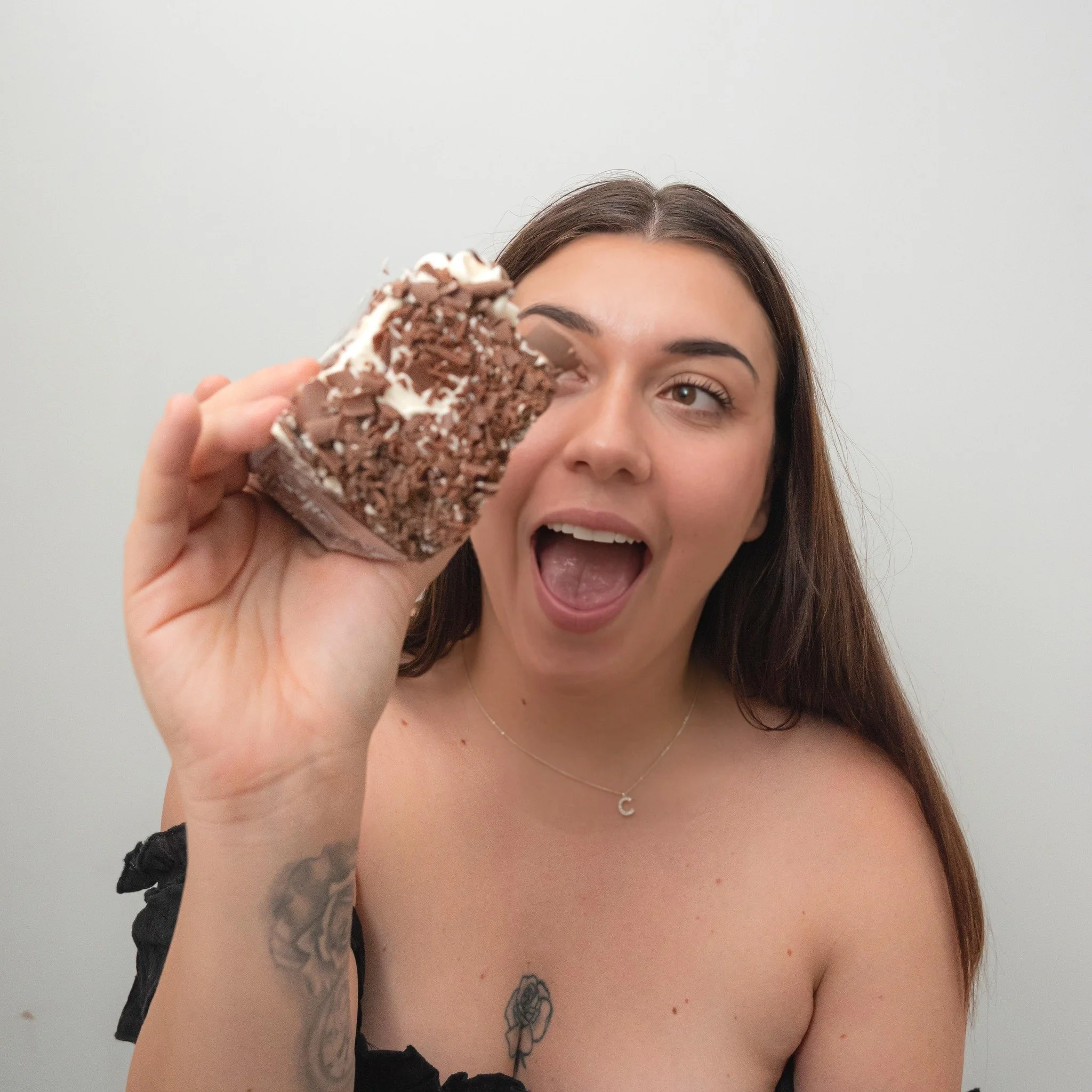 A woman with long brown hair and a tattoo of a rose on her chest holds a large chocolate dessert near her face, smiling and sticking her tongue out, against a plain white background.