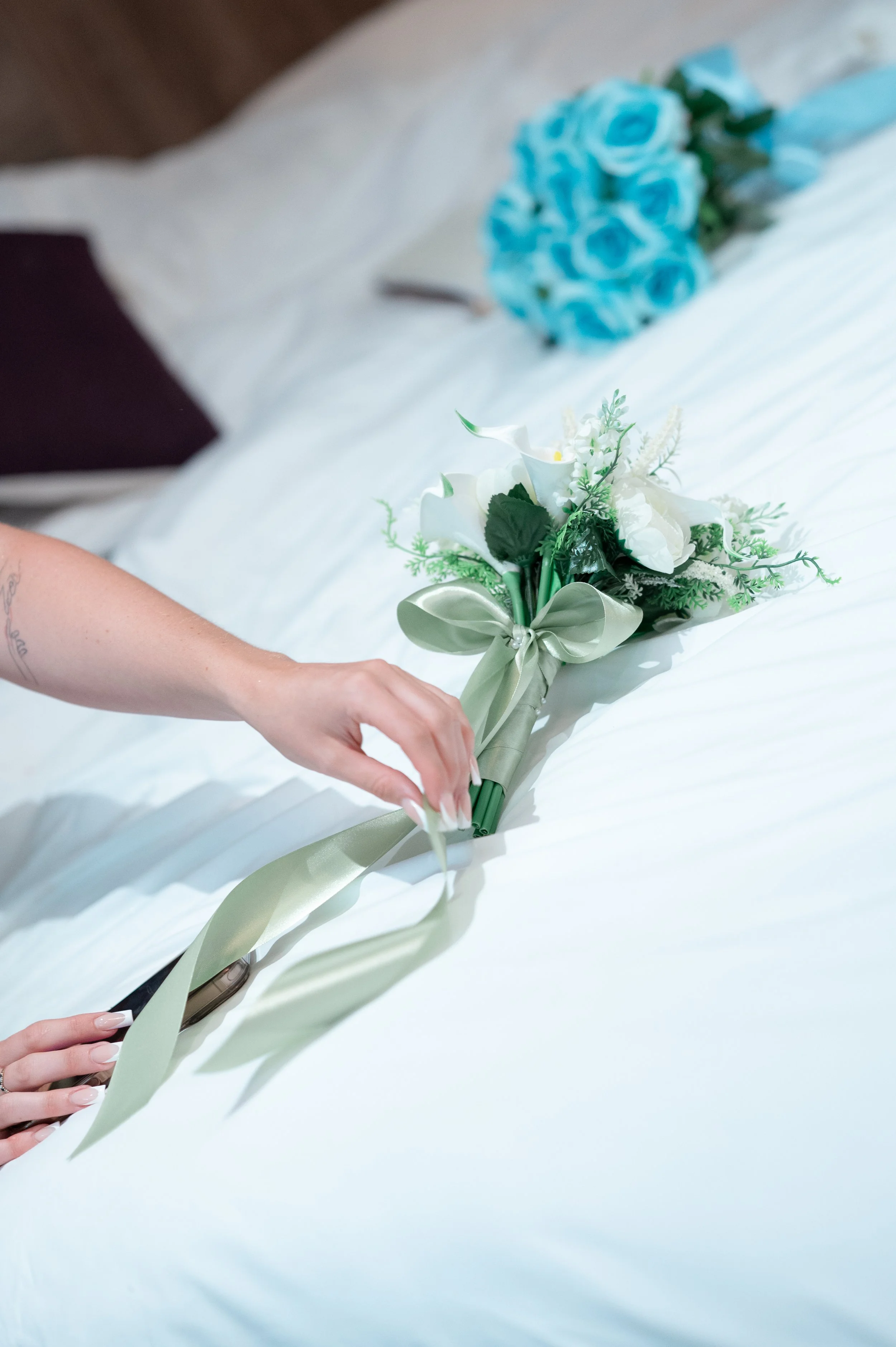 Person holding a bouquet of white flowers with green leaves and a pale ribbon, on a white surface. In the background, there is a bundle of blue roses.