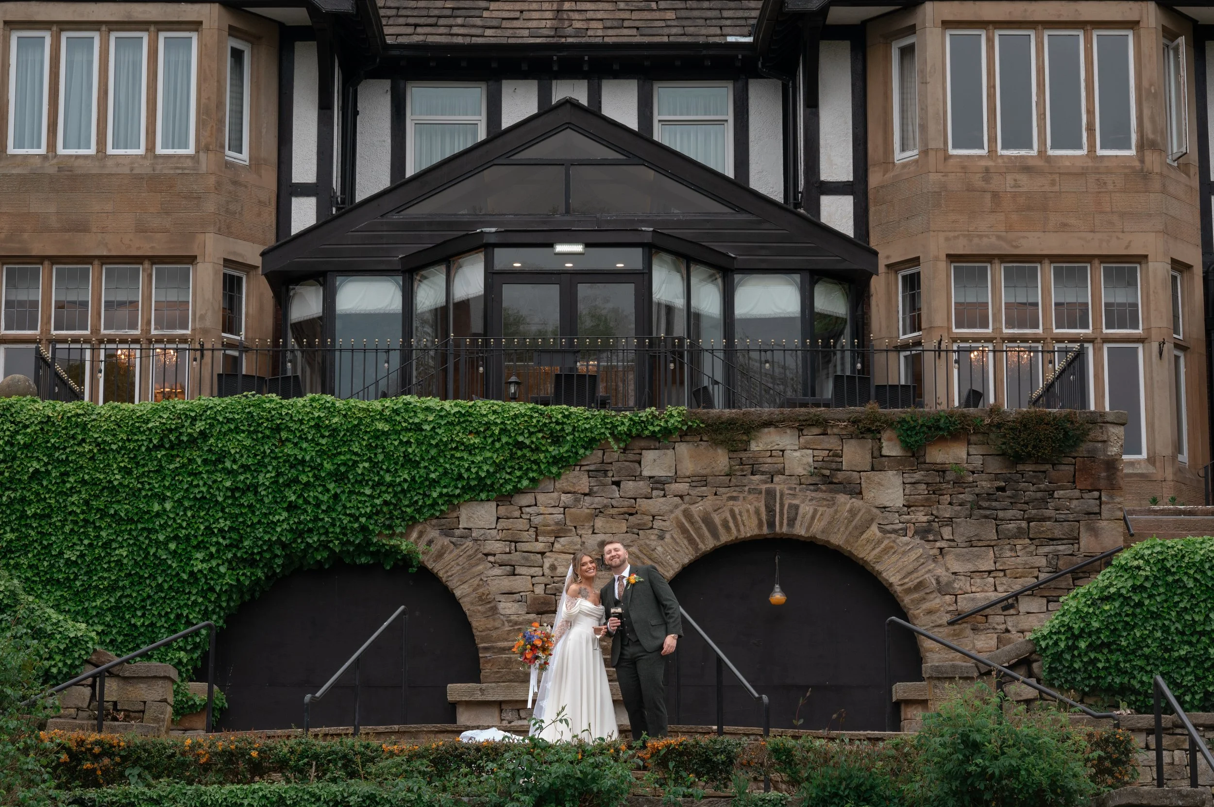 A bride and groom standing on a staircase in front of a building with multiple windows, a stone arch, and greenery. The bride is in a white wedding dress holding a bouquet of colorful flowers, and the groom is in a dark suit with a bow tie.