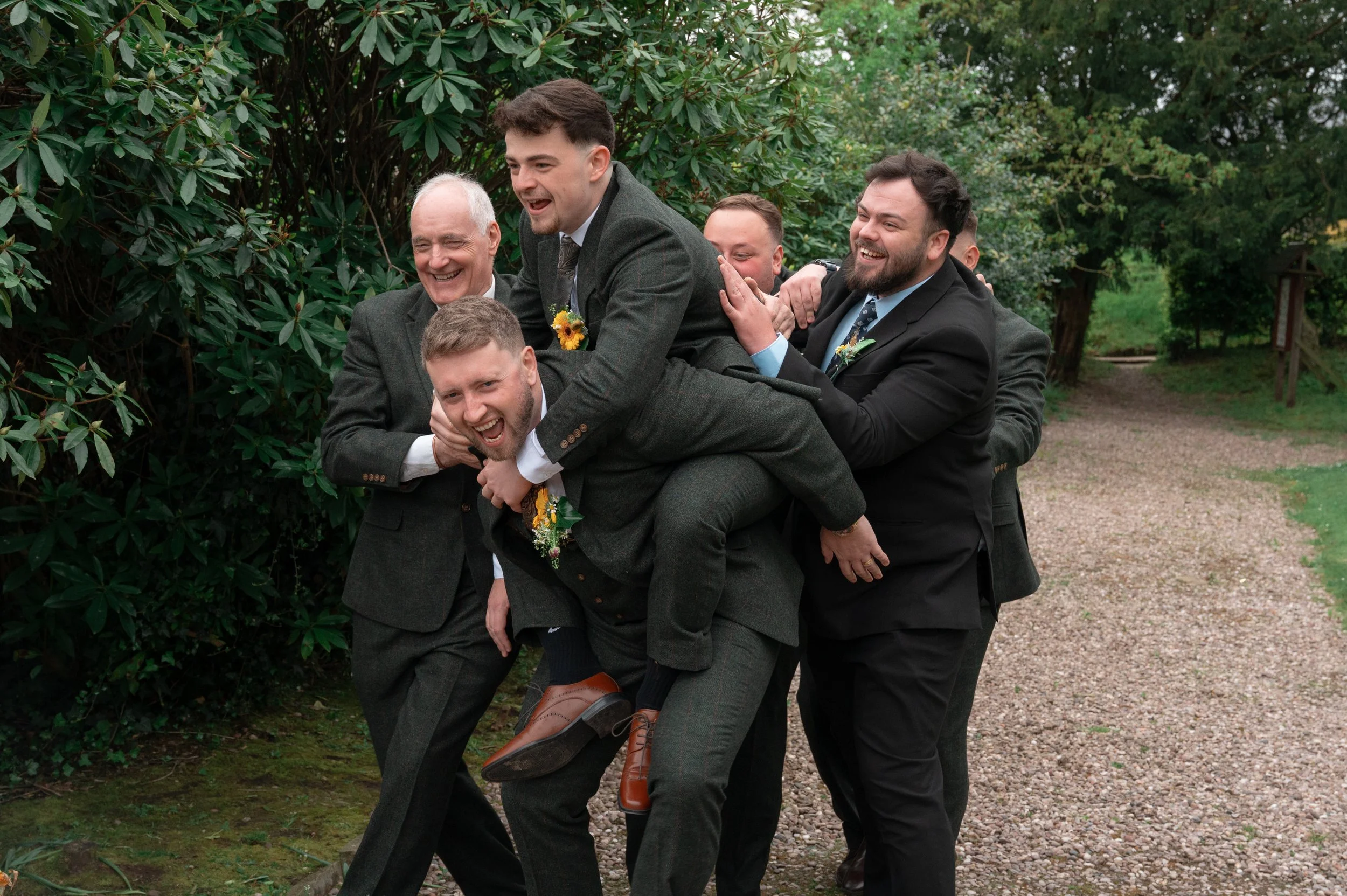 A group of men dressed in suits outdoors, playing a game of conga line, smiling and laughing, with lush green trees in the background.