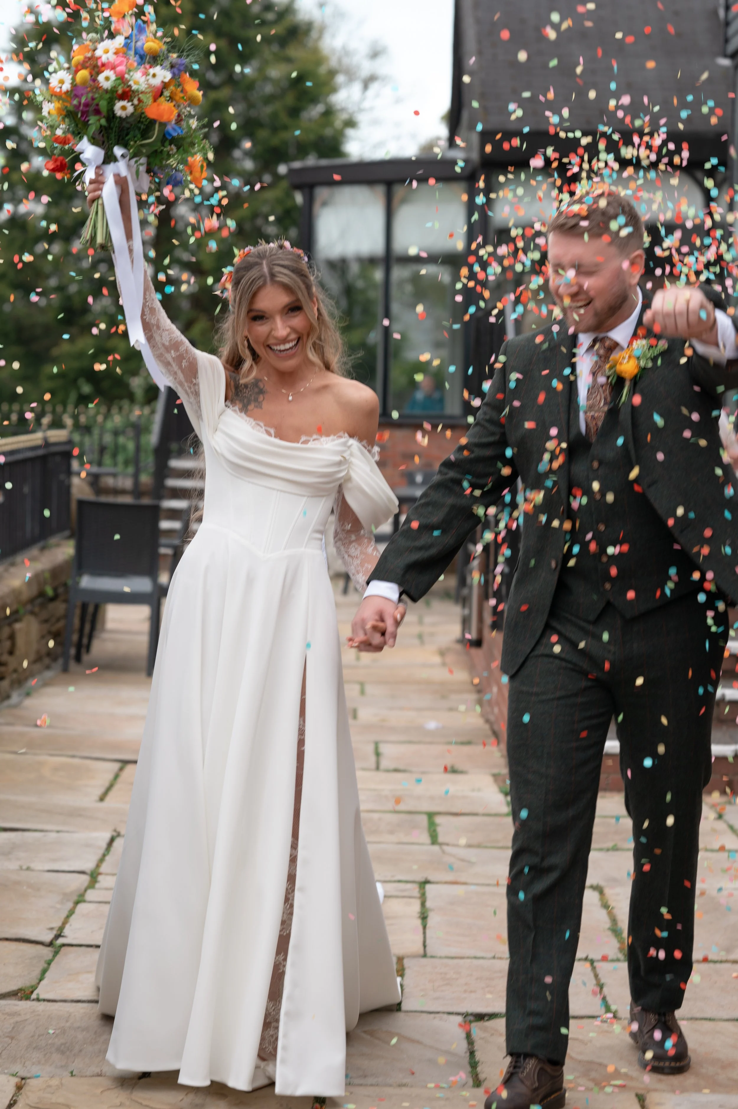 A bride and groom hold hands and smile as they celebrate their wedding outdoors. The bride holds a bouquet of colorful flowers raised in the air, while confetti falls around them. The bride is dressed in a white wedding gown, and the groom is wearing