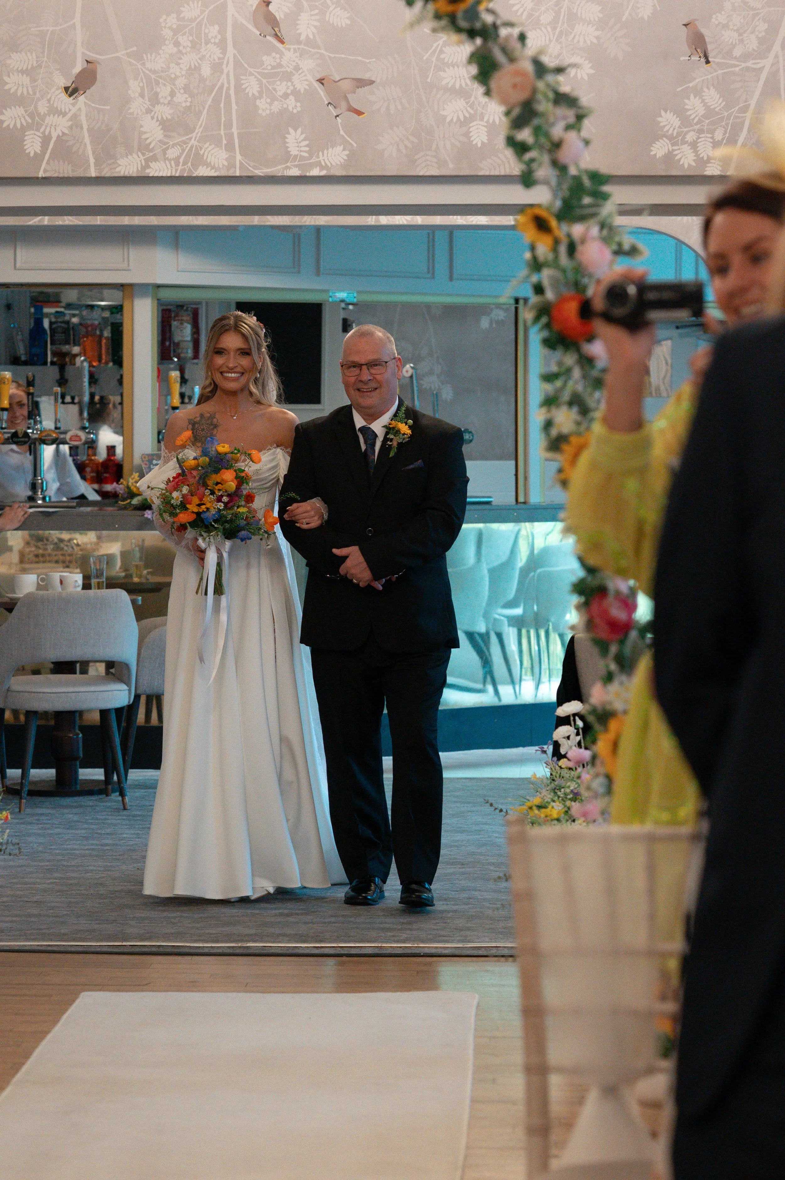 A bride walking down the aisle with her father during a wedding ceremony. The bride is smiling and holding a colorful bouquet of flowers. The background shows a decorated wedding venue with floral arrangements and a wall with a bird pattern.