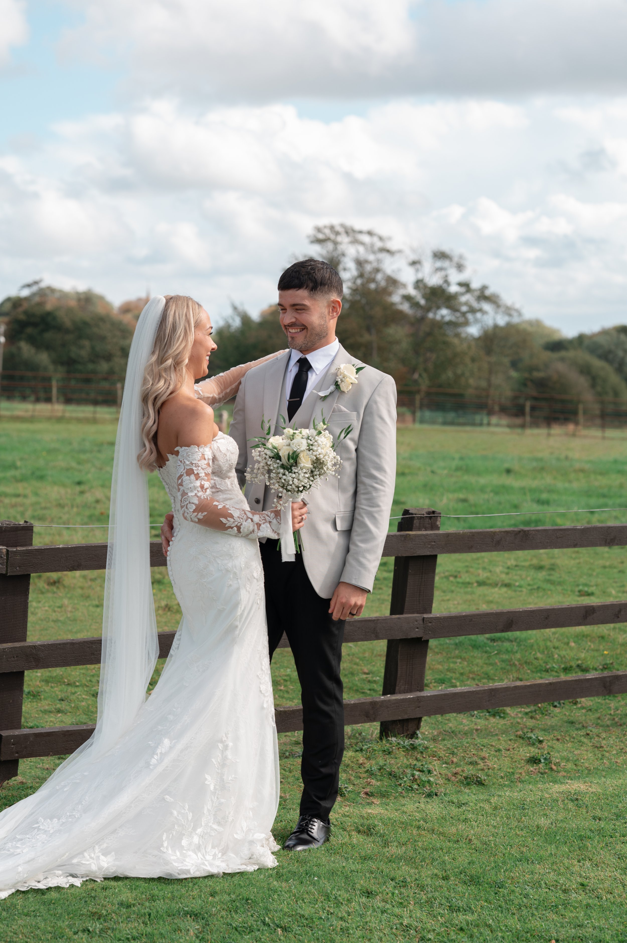A bride and groom smiling at each other outdoors on their wedding day, with the bride holding a bouquet of white flowers, standing behind a wooden fence, with a grassy field and scattered trees in the background under a partly cloudy sky.