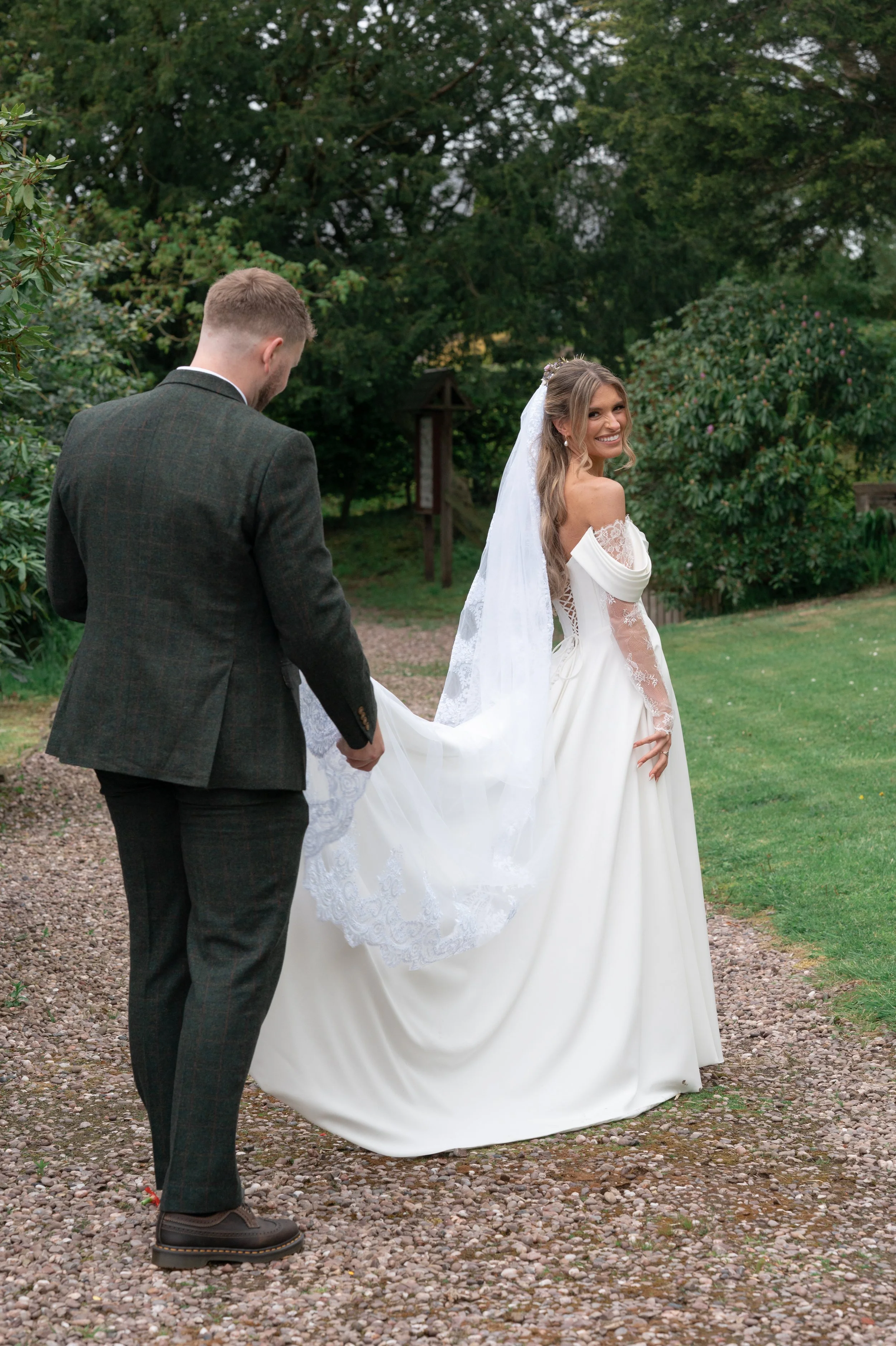 A bride in a white wedding gown smiling and looking over her shoulder, holding the train of her dress, as a groom in a dark suit stands nearby in an outdoor garden setting with trees and greenery.