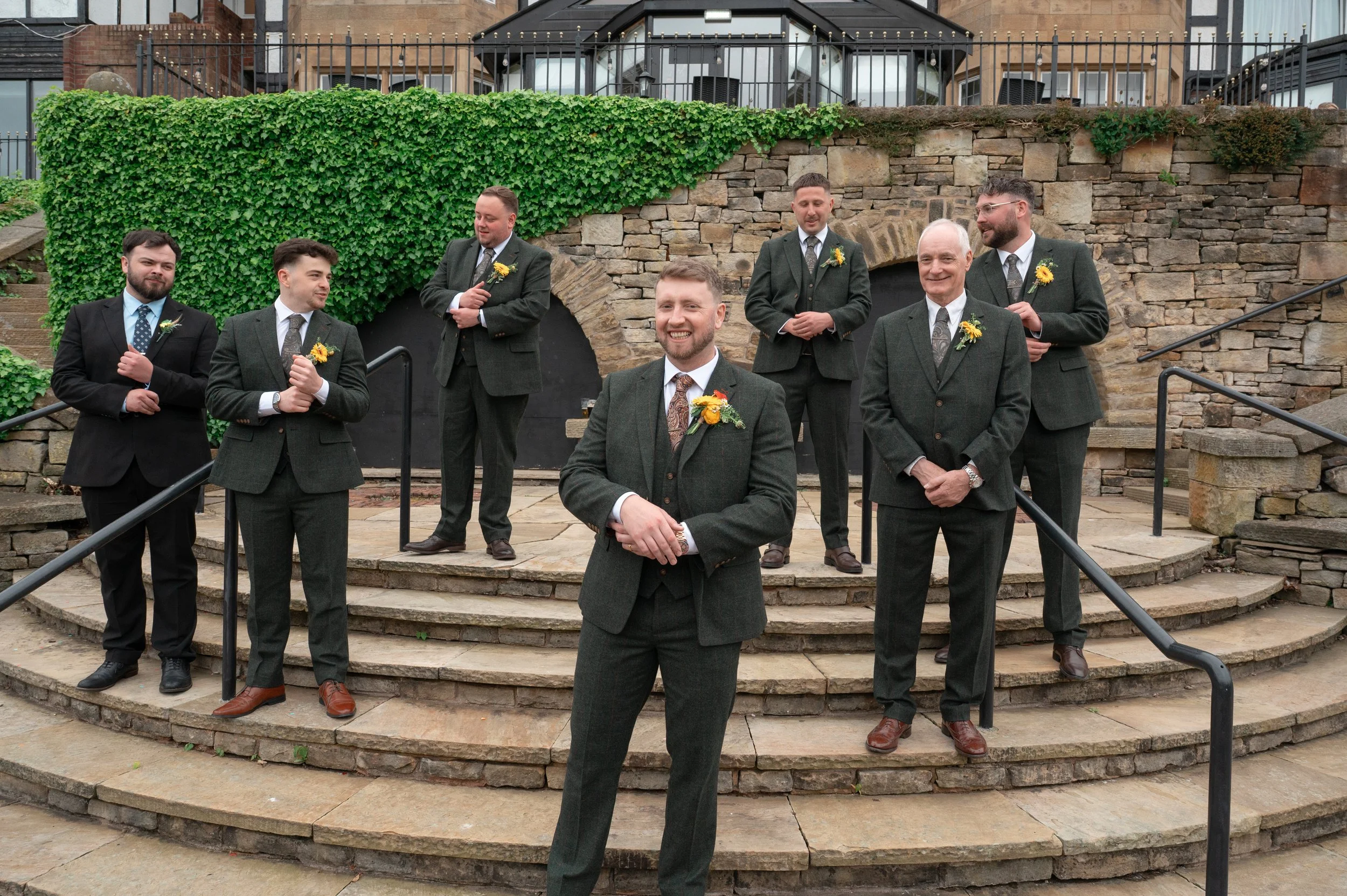 Seven men dressed in suits with boutonnières, standing on stone steps outside, smiling and chatting at a formal event.