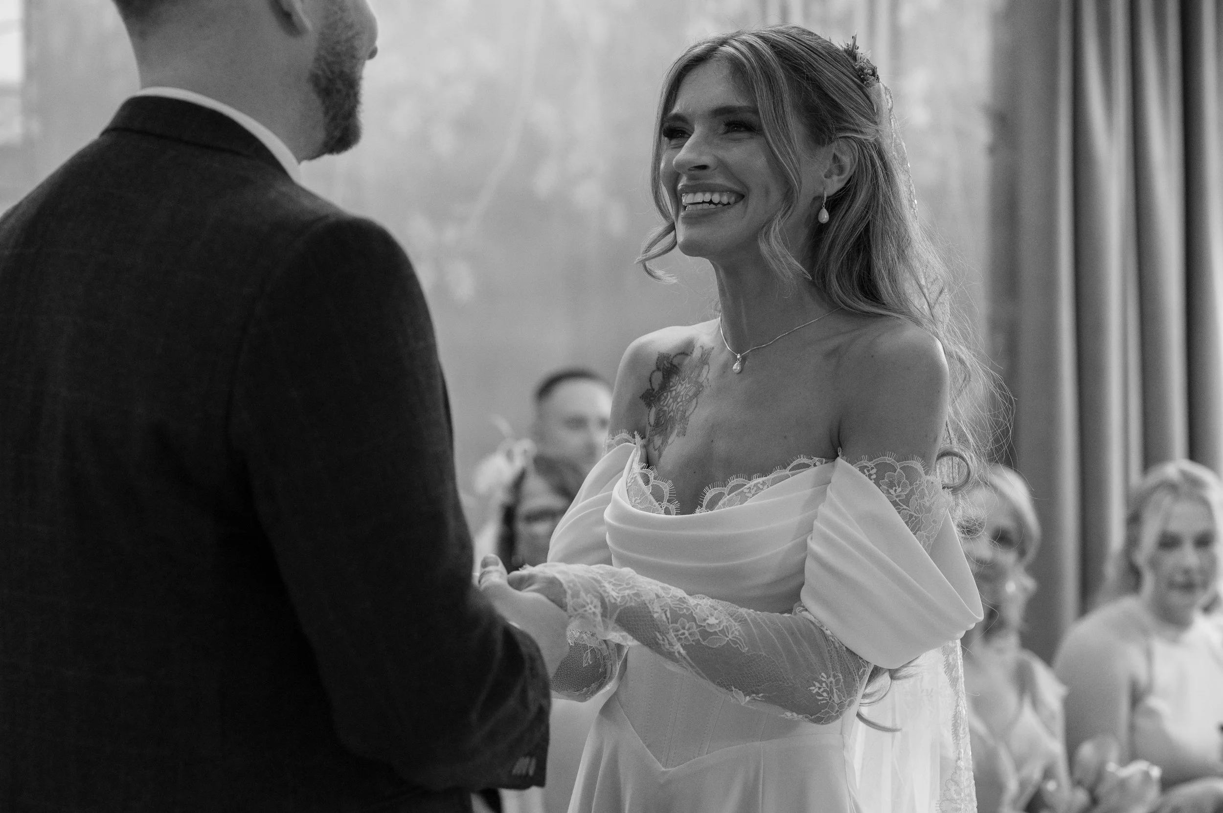 A bride with long wavy hair wearing a bridal gown with lace details, smiling at her groom during their wedding ceremony.