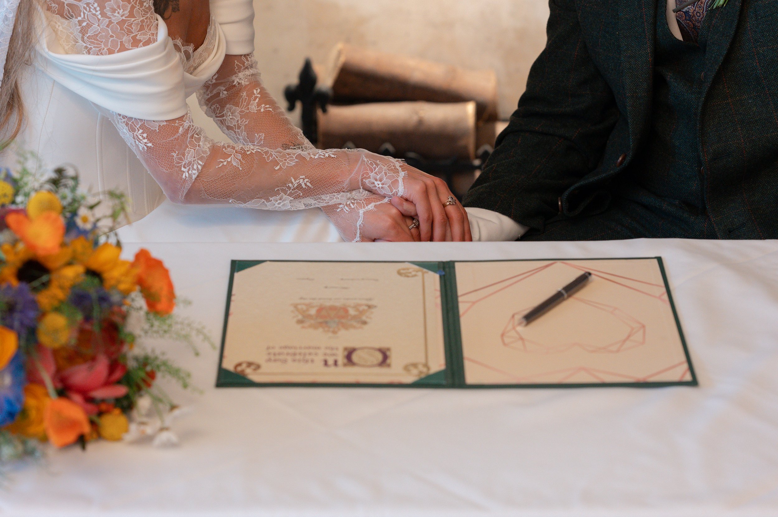 A wedding ceremony with a bride and groom holding hands over a table with paperwork, pens, and a bouquet of colorful flowers in the foreground.