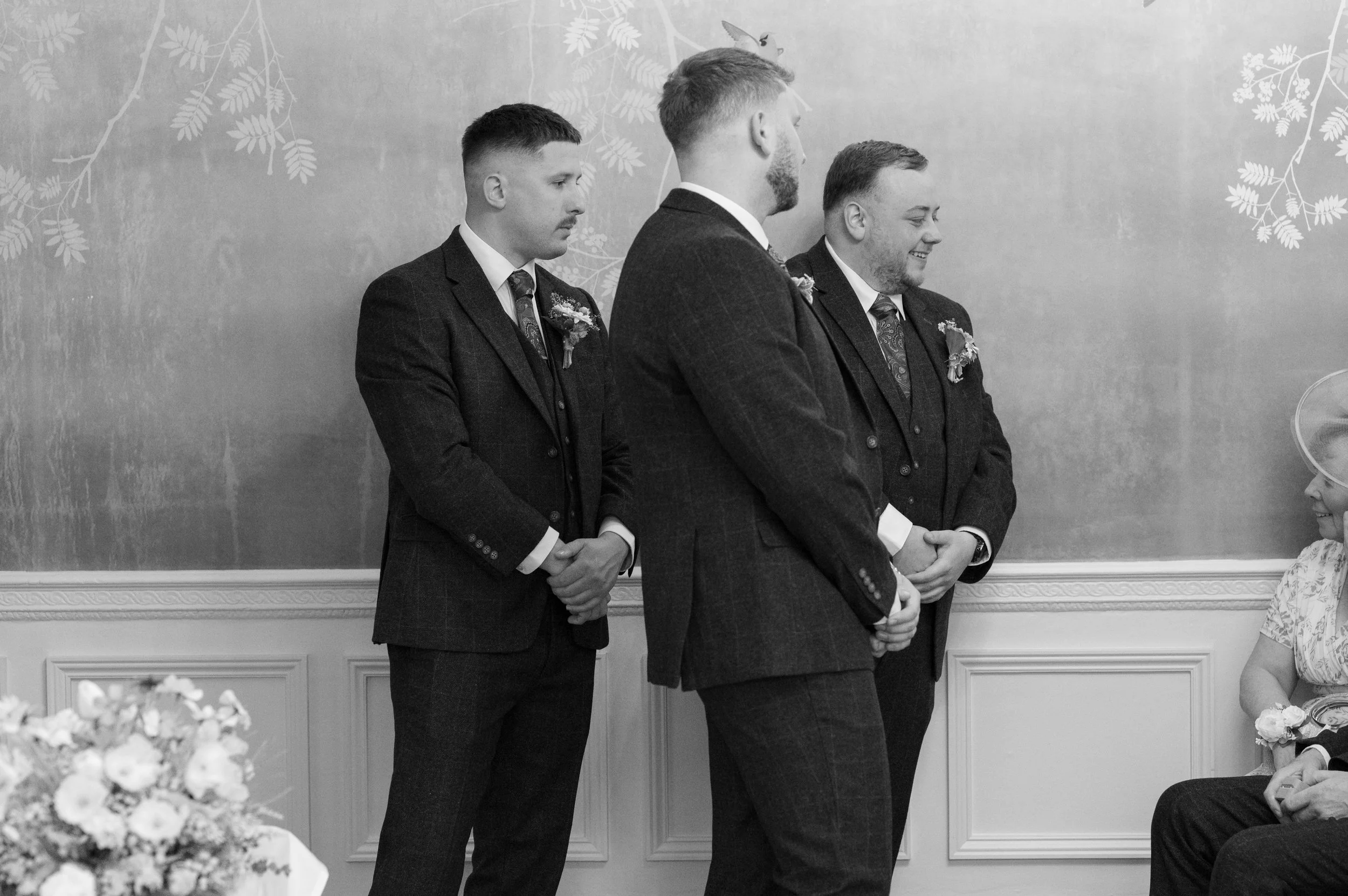 Four men in suits standing in a line, holding hands, smiling, and participating in a wedding ceremony. An older woman is seated to the right, smiling at the event. The background features a decorative wall with a floral and bird pattern, and a table 