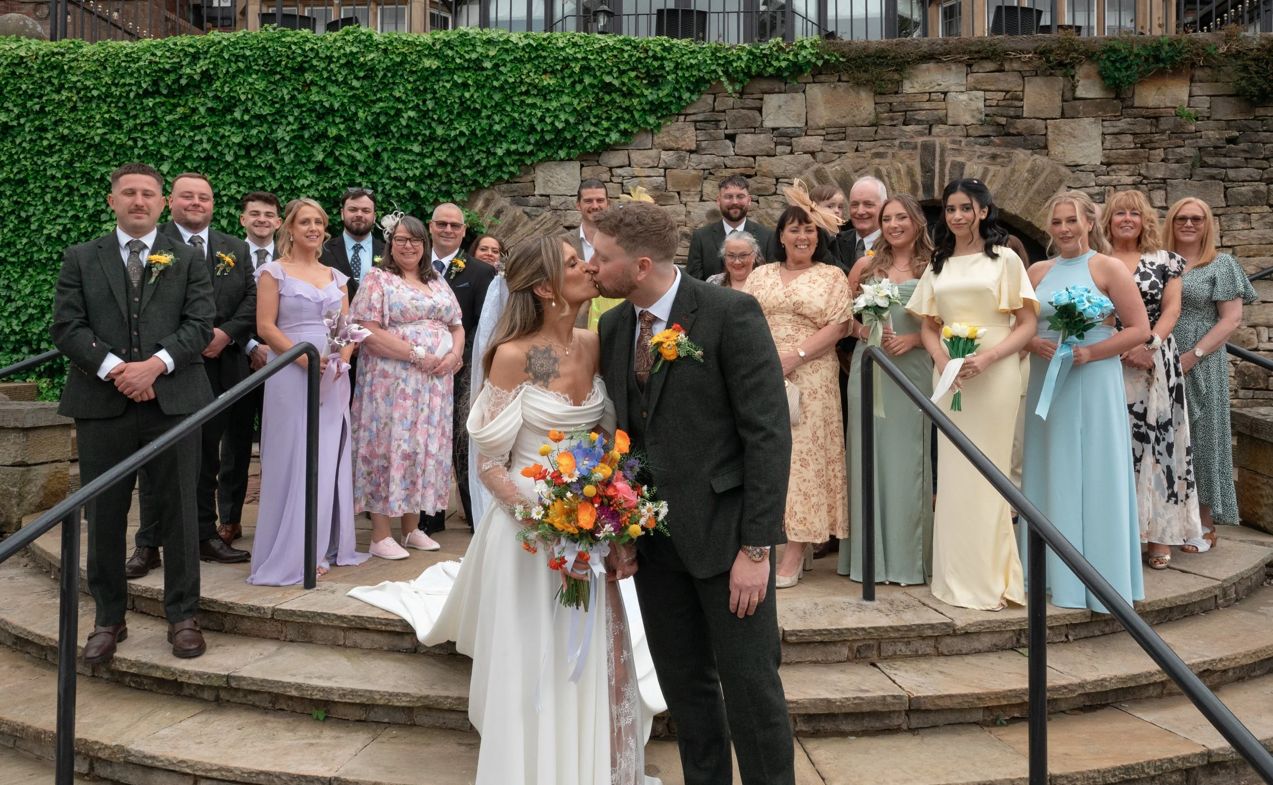 A bride and groom share a kiss during their wedding ceremony on stone steps, surrounded by friends and family in formal attire.