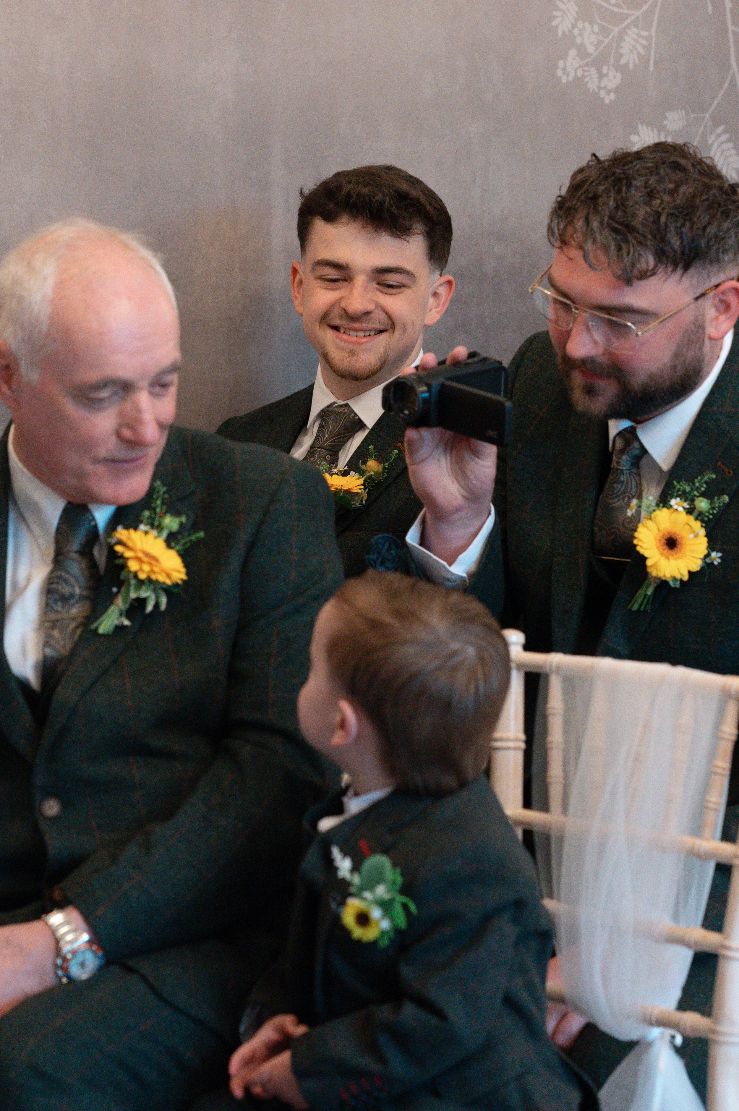 A group of men and boys dressed in formal attire, sitting closely together at an event, with some of them wearing boutonnières and floral accessories, engaged in conversation and smiling.