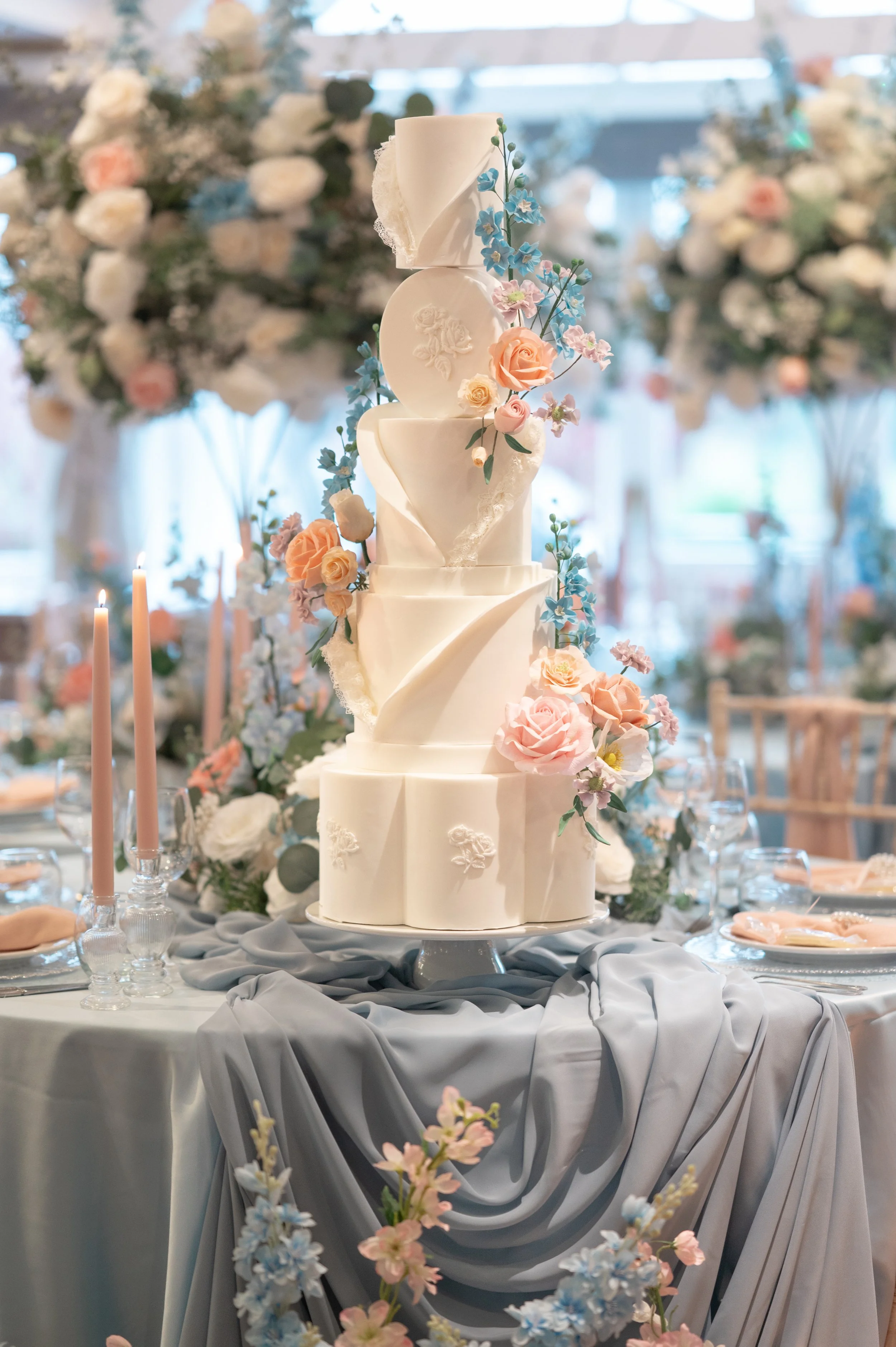 Elegant white wedding cake with floral decorations on a table with gray draped fabric, surrounded by pink and blue flowers.