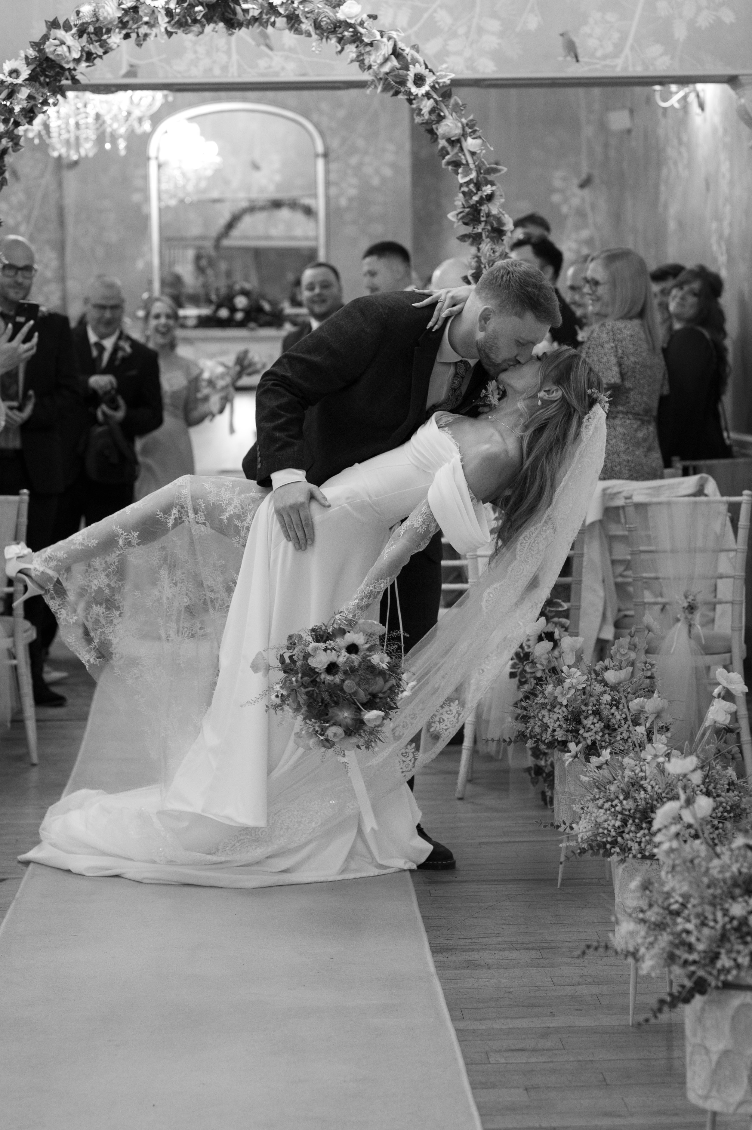 A wedding ceremony showing a groom leaning over and kissing a bride who is lying on a table covered with a lace cloth, surrounded by floral arrangements, with guests watching and taking photos in the background.