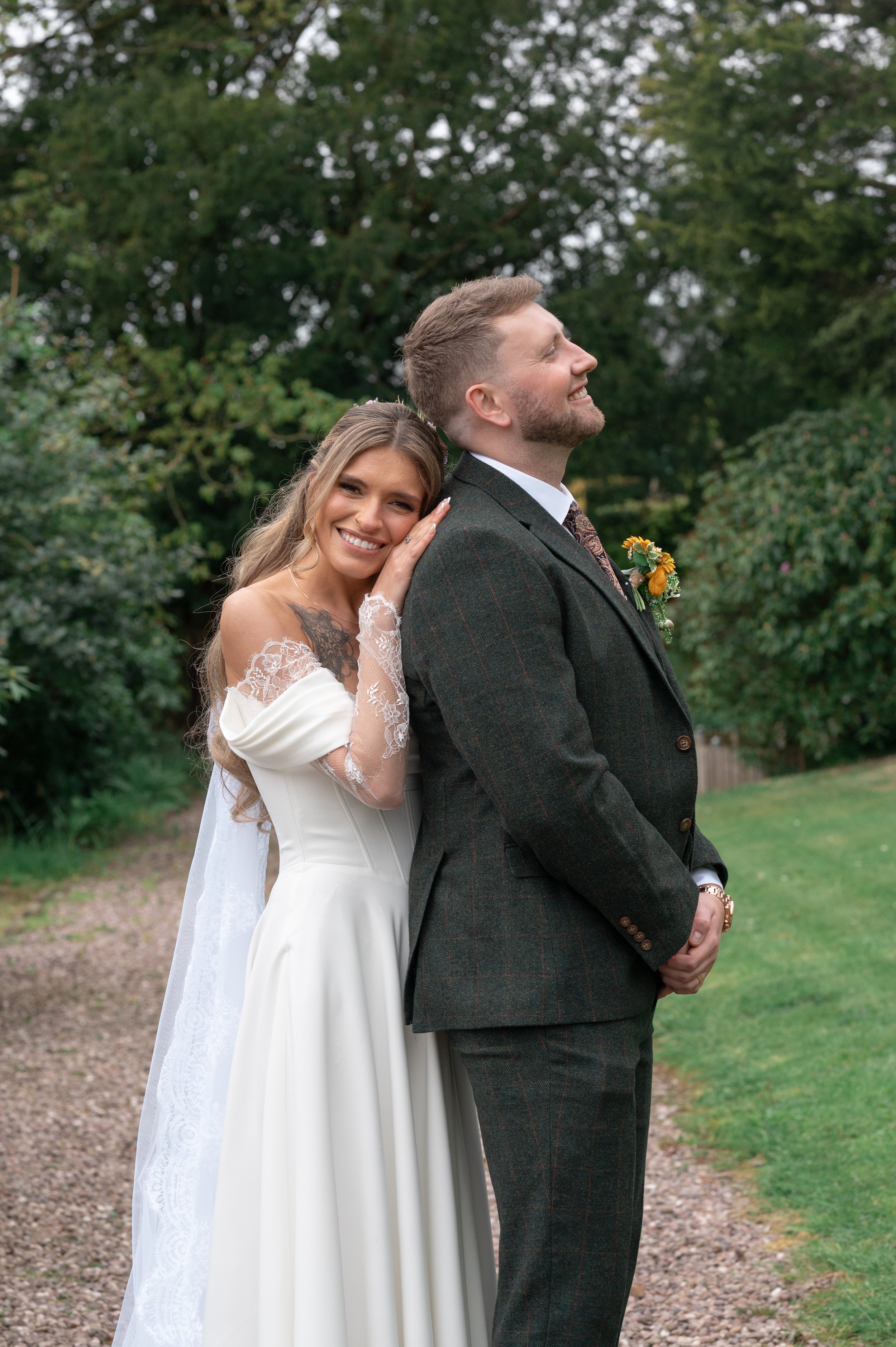 A bride and groom stand outside in a green park during daytime. The bride leans on the groom's back with a smile, wearing a white wedding dress with lace details on the sleeves. The groom is dressed in a dark, checked suit with a white shirt and a pa
