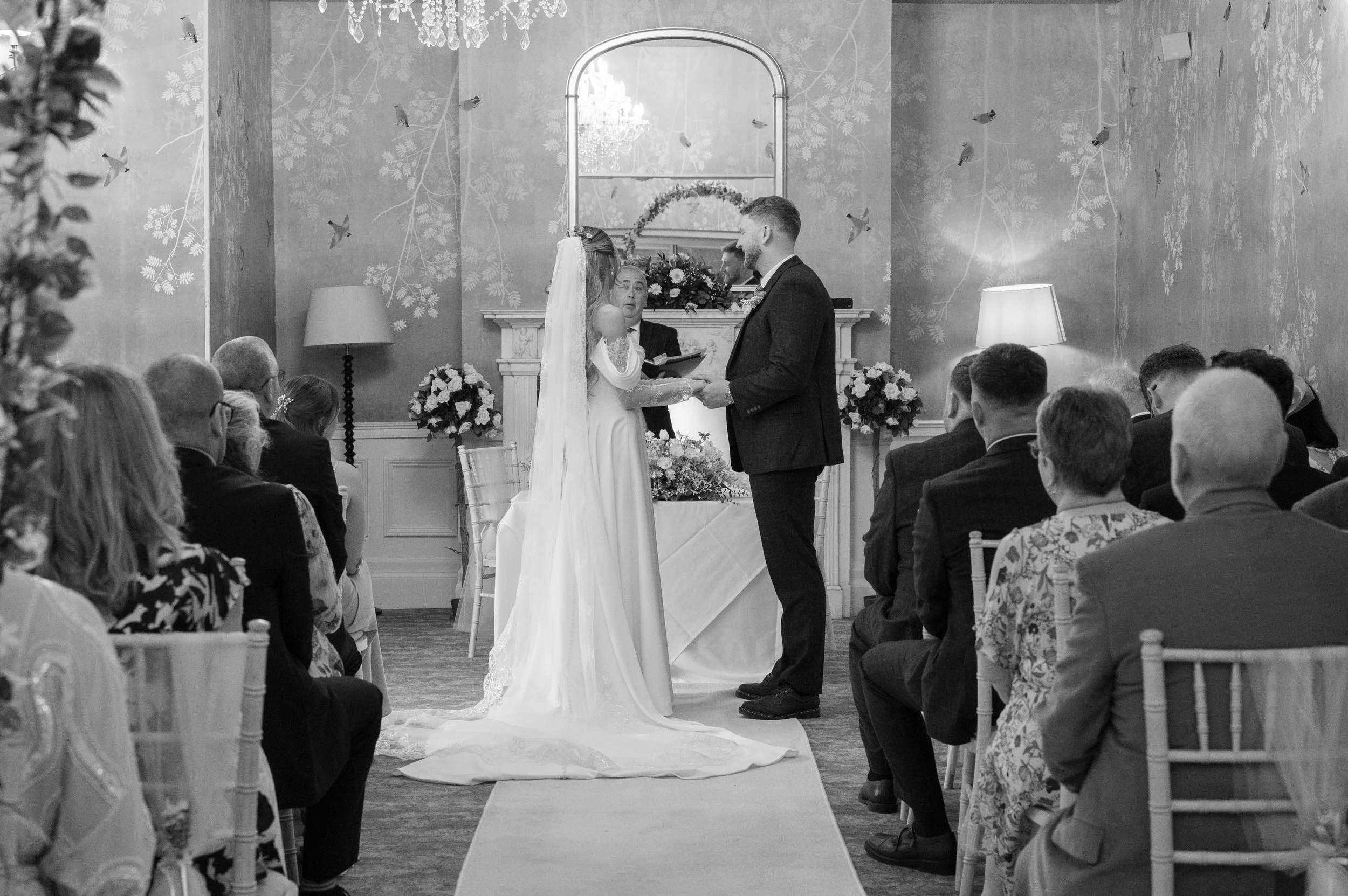 A black and white photo of a wedding ceremony with a bride and groom standing face to face, exchanging vows in front of an officiant. Guests are seated on chairs facing them, and the room is decorated with floral arrangements, wall art, and a chandel