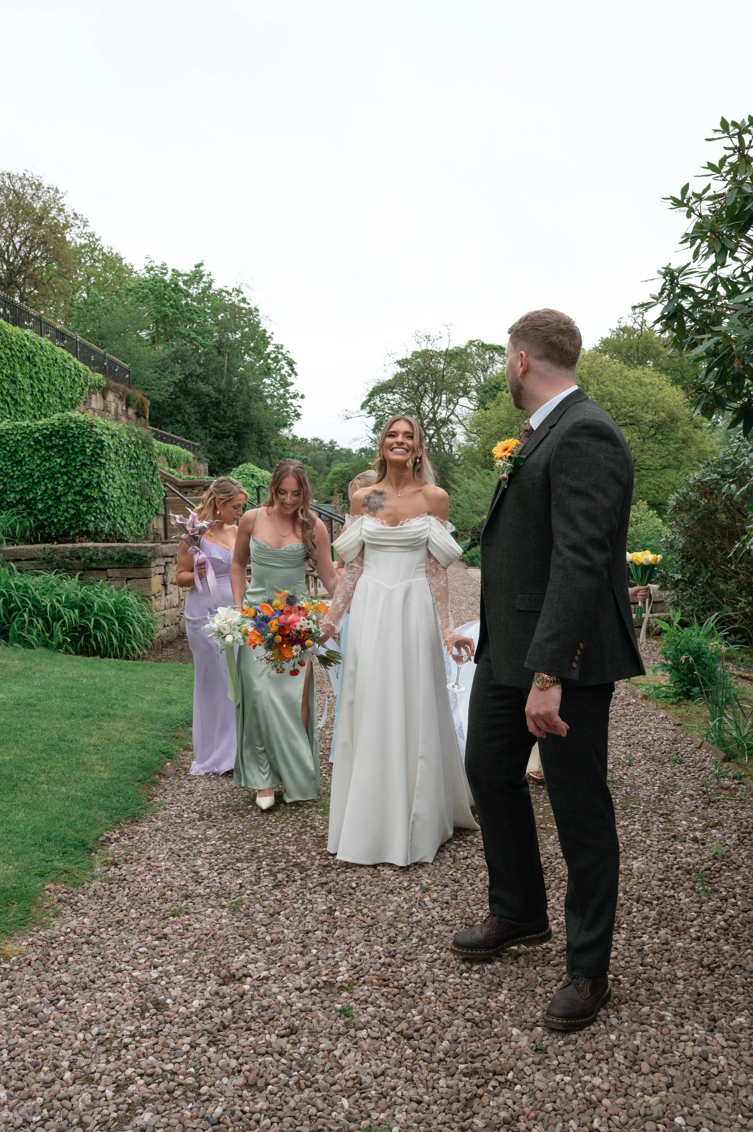 A bride in a white wedding dress with off-shoulder sleeves holding a colorful bouquet, smiling at and holding hands with a groom in a dark suit with a sunflower boutonniere, accompanied by bridesmaids in pastel dresses, outdoors in a garden with gree