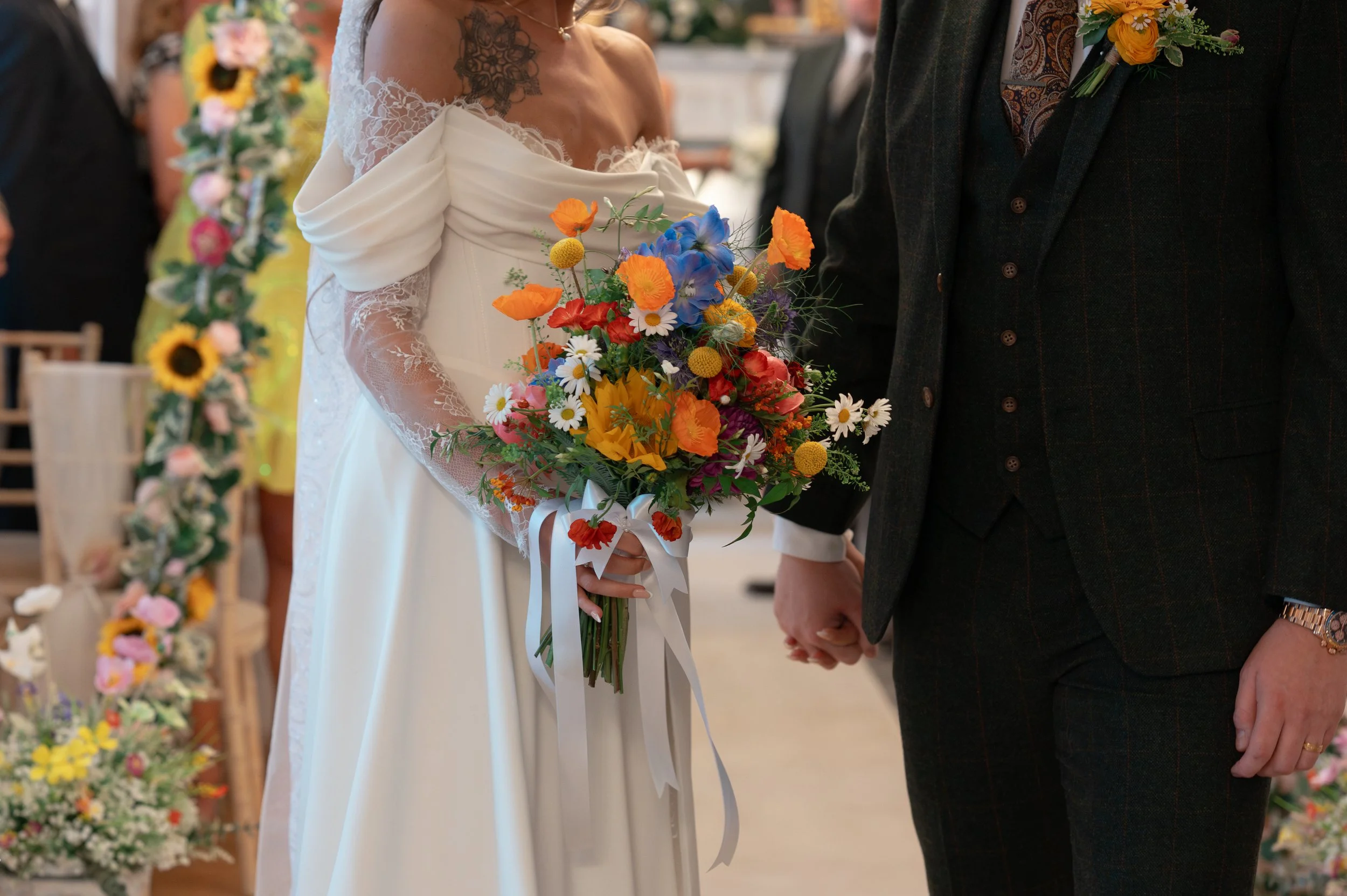 A bride in a white wedding dress holding a colorful bouquet of flowers, standing next to a groom dressed in a dark suit, both holding hands during a wedding ceremony.