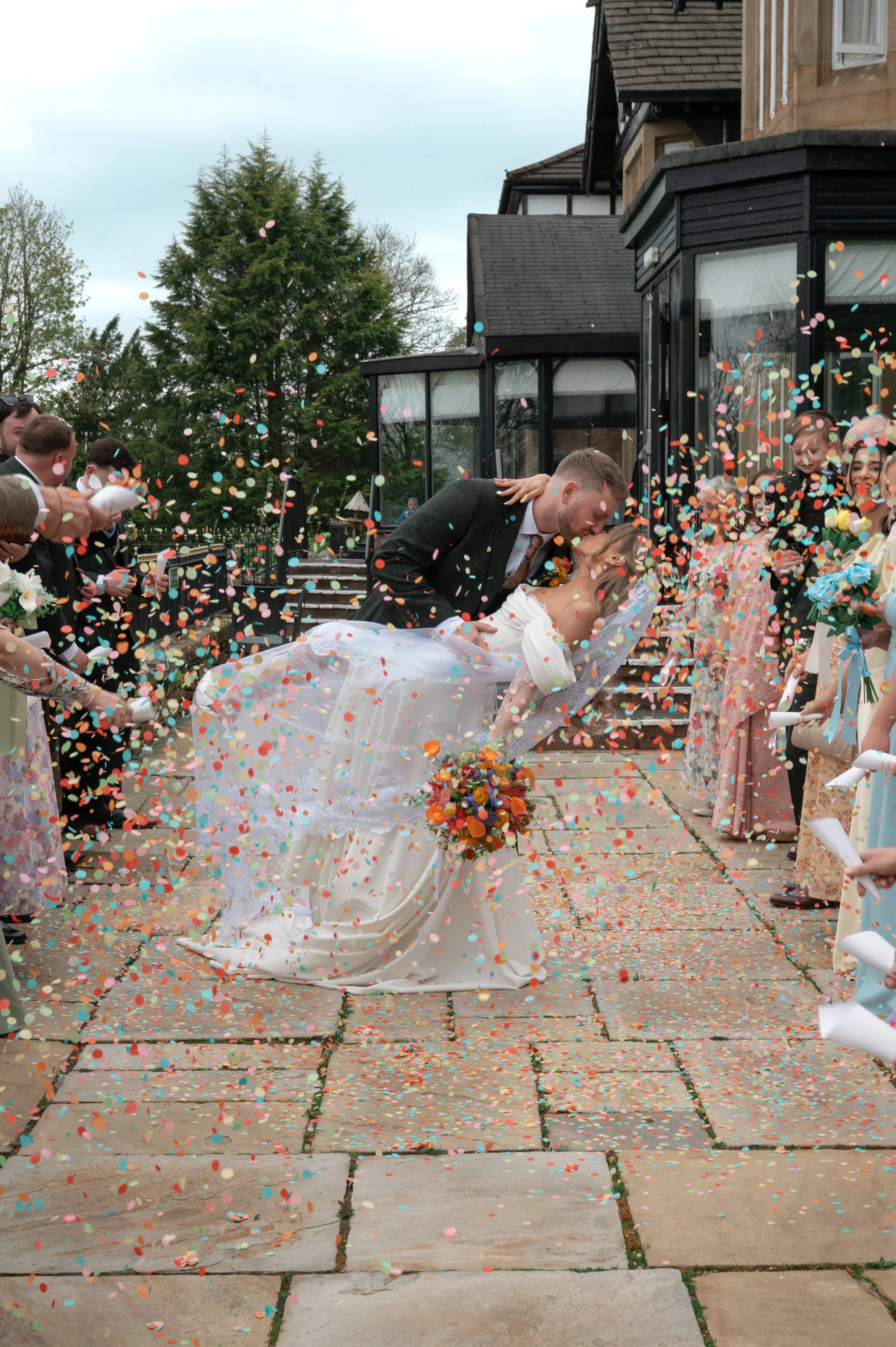 A bride and groom share a kiss during their wedding celebration, surrounded by confetti and guests.