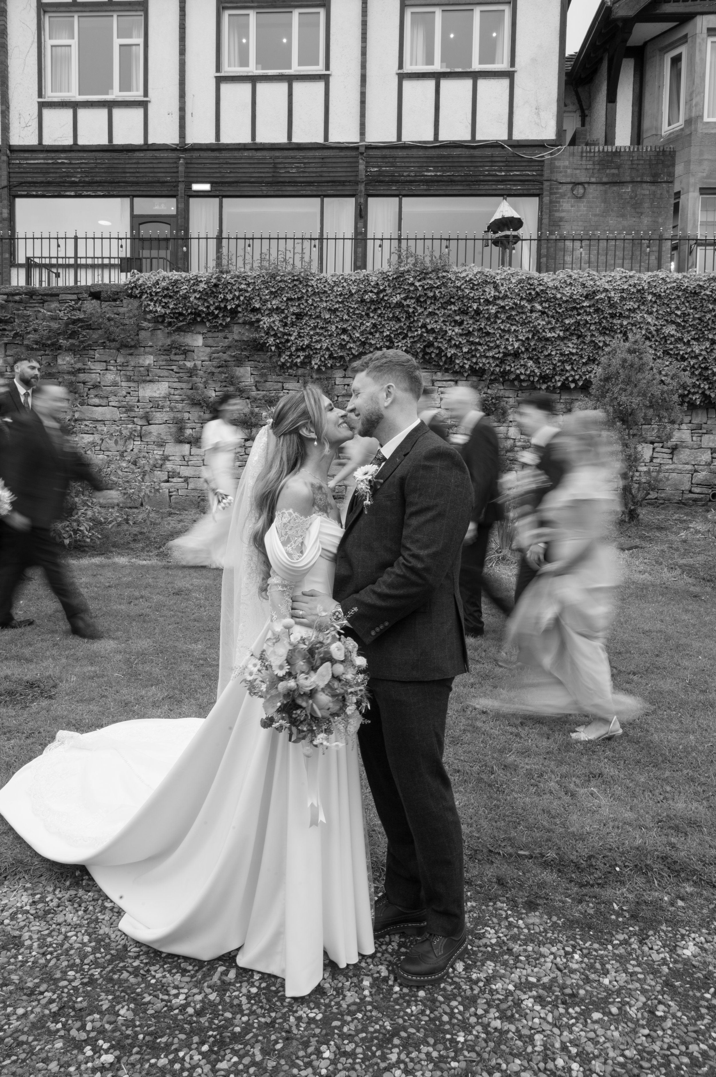 A wedding couple stands close together, smiling and looking into each other's eyes, surrounded by blurred guests walking past them at an outdoor wedding ceremony.