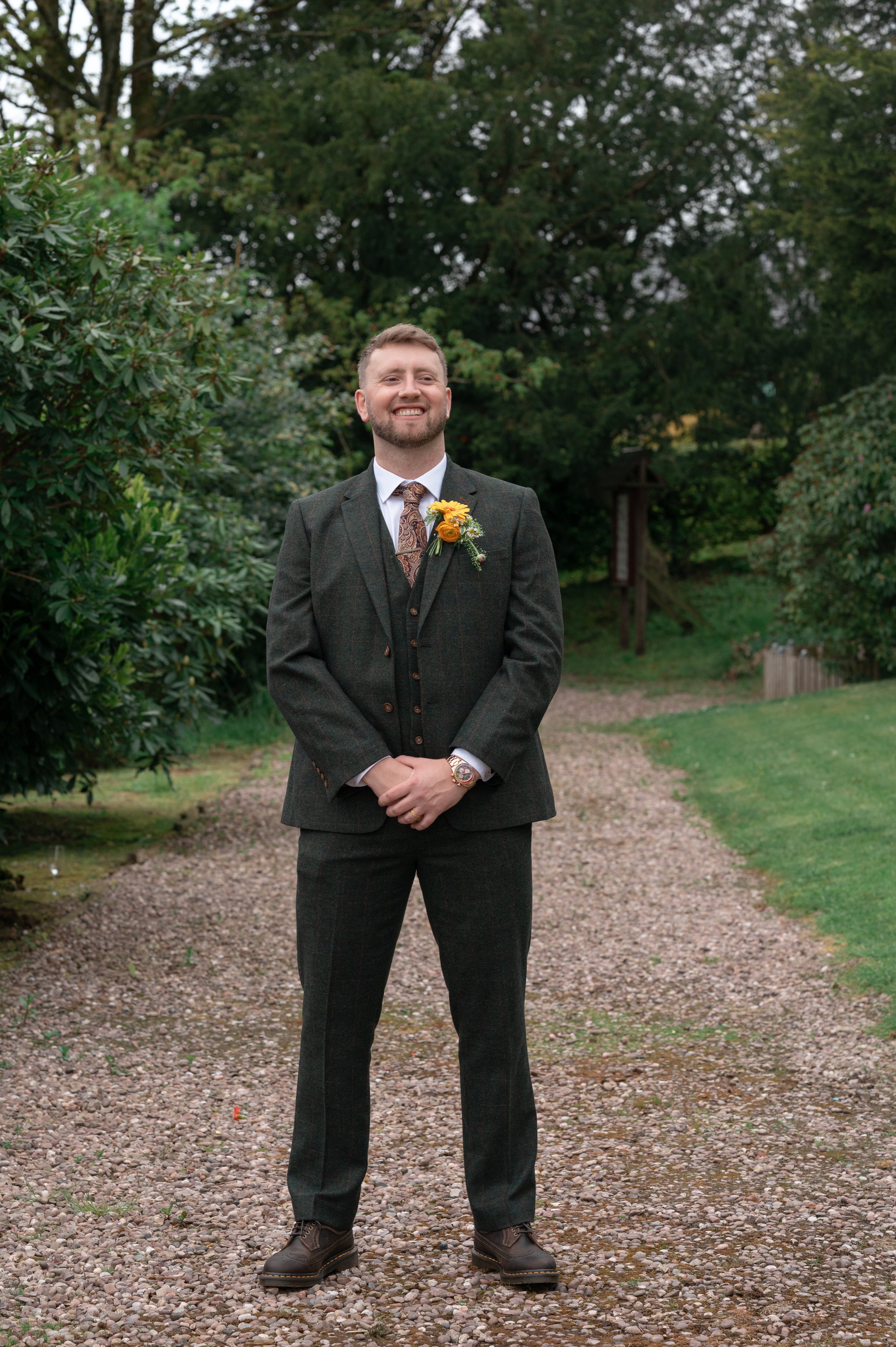 A man in a suit standing outdoors on a gravel path surrounded by greenery, smiling.