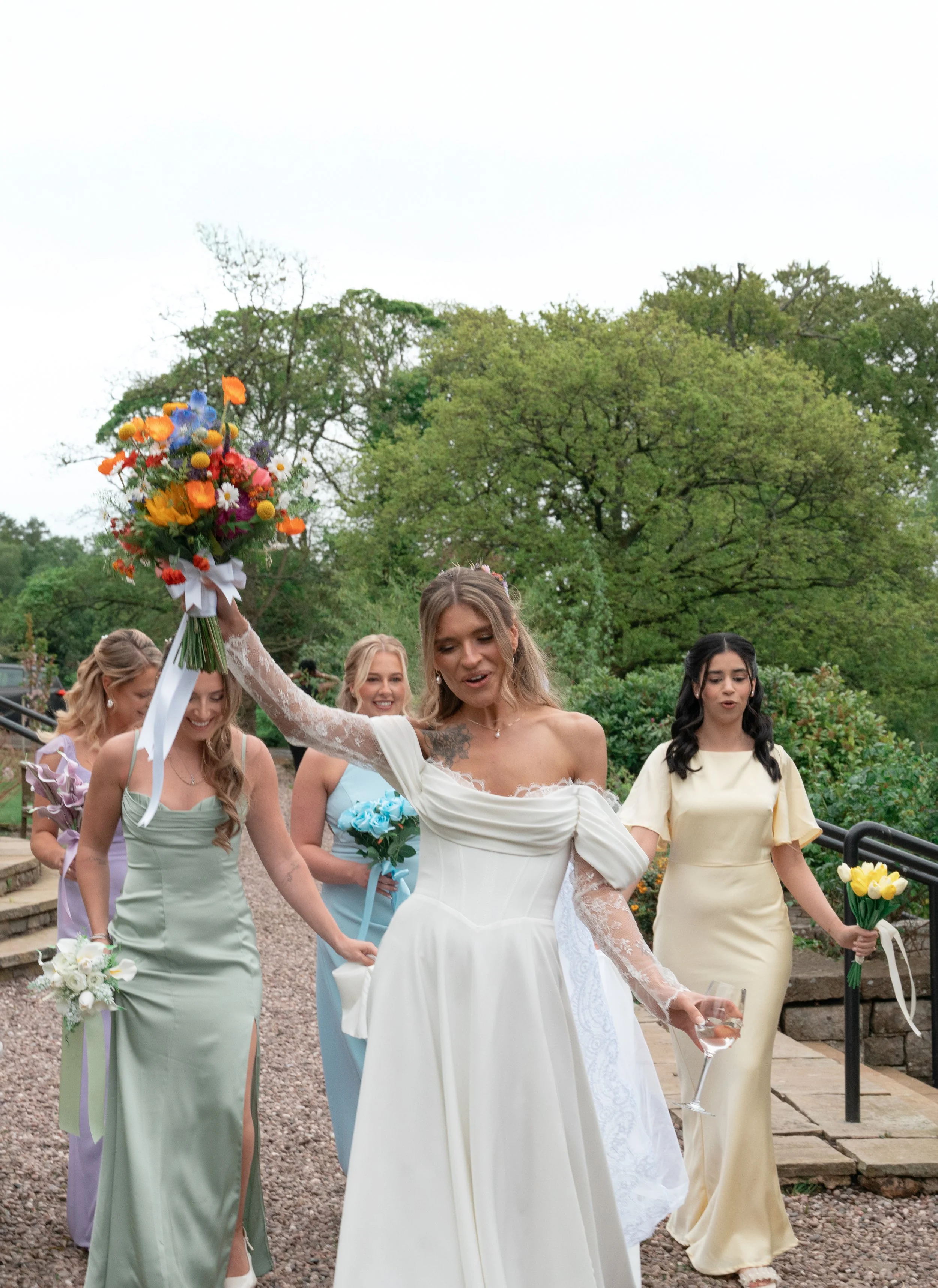 Women in pastel dresses celebrating outdoors, with one holding a bouquet of flowers and another holding a glass of wine, surrounded by greenery and trees.