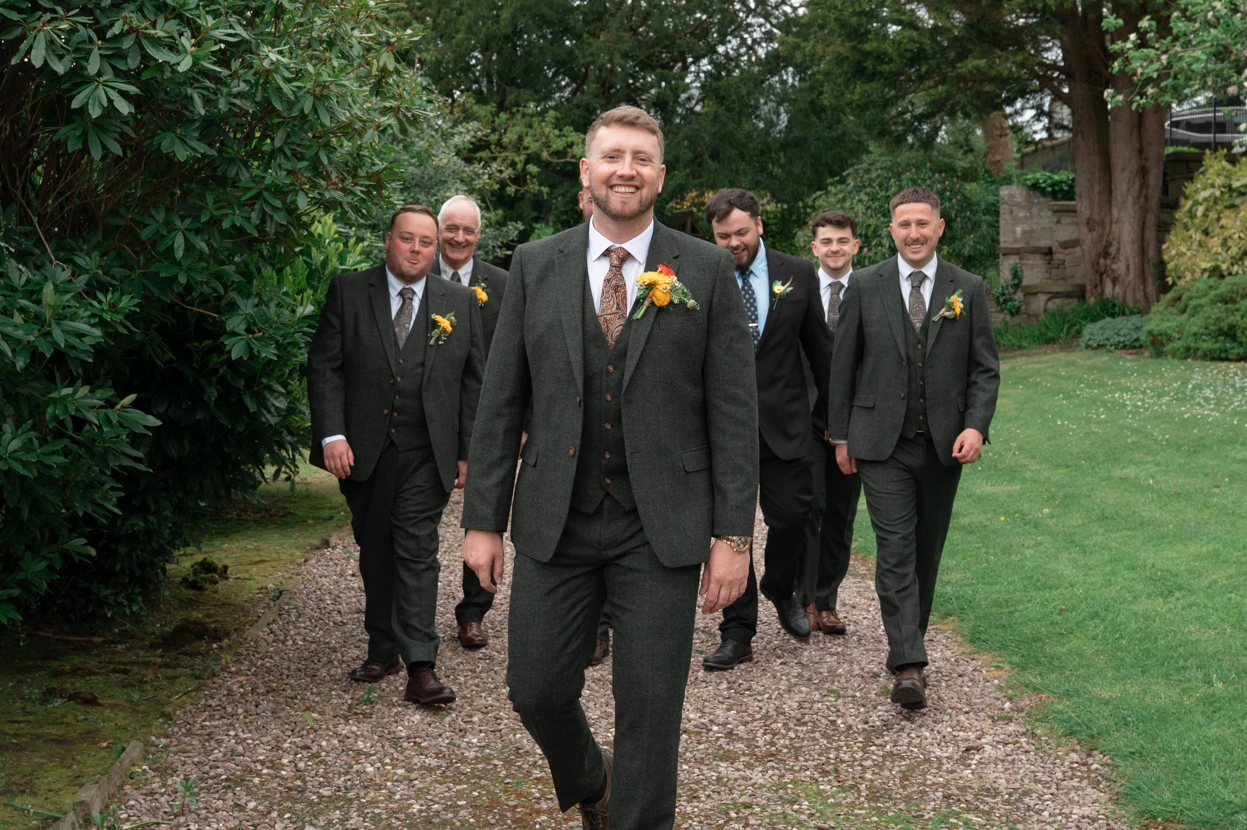 A groom and six men in suits walking outdoors on a garden pathway, smiling, with the groom leading.