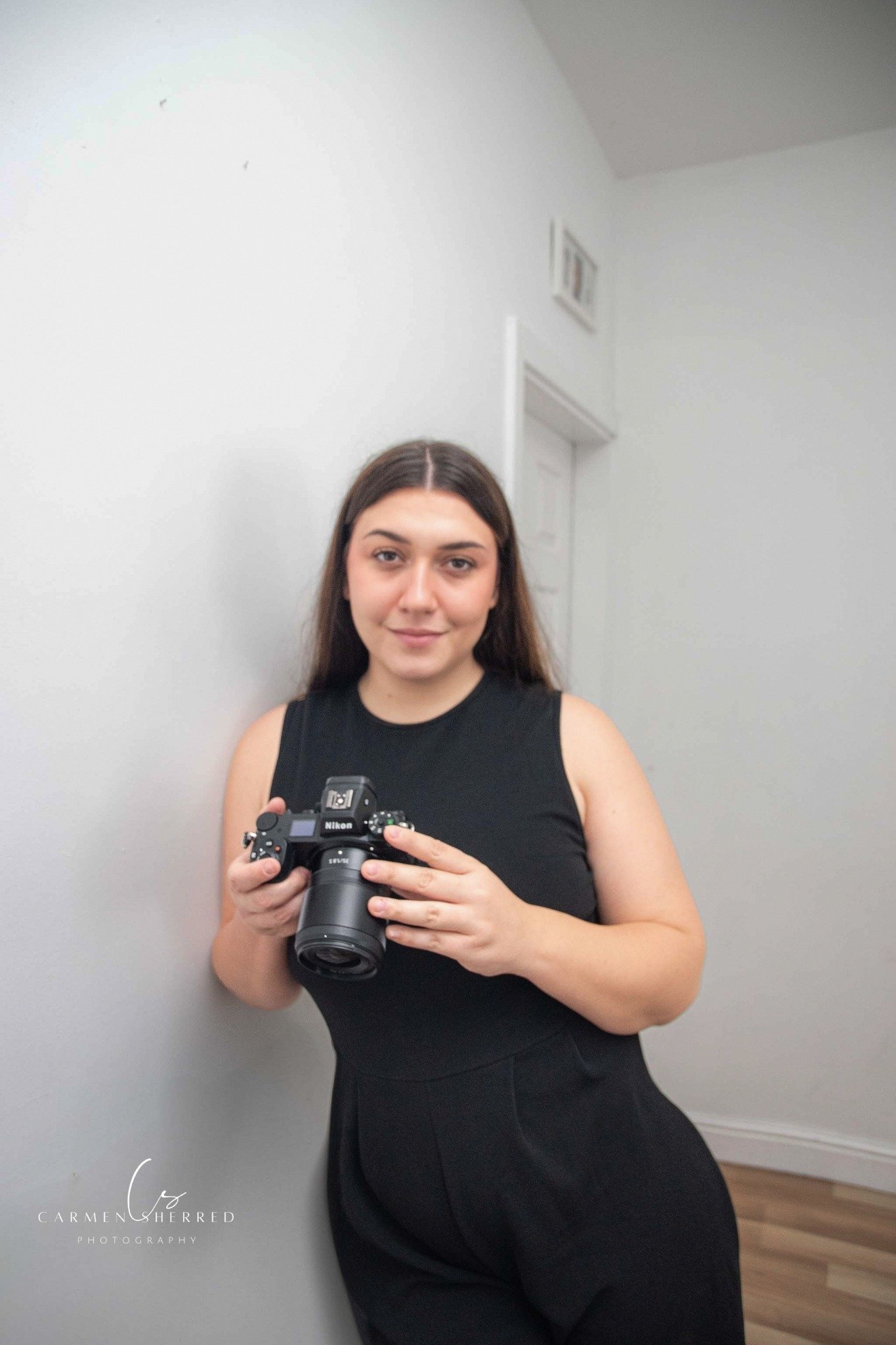 A woman with long brown hair taking a mirror selfie with a DSLR camera. She is wearing a black sleeveless top and black pants, standing against a plain white wall in a room with wooden flooring.