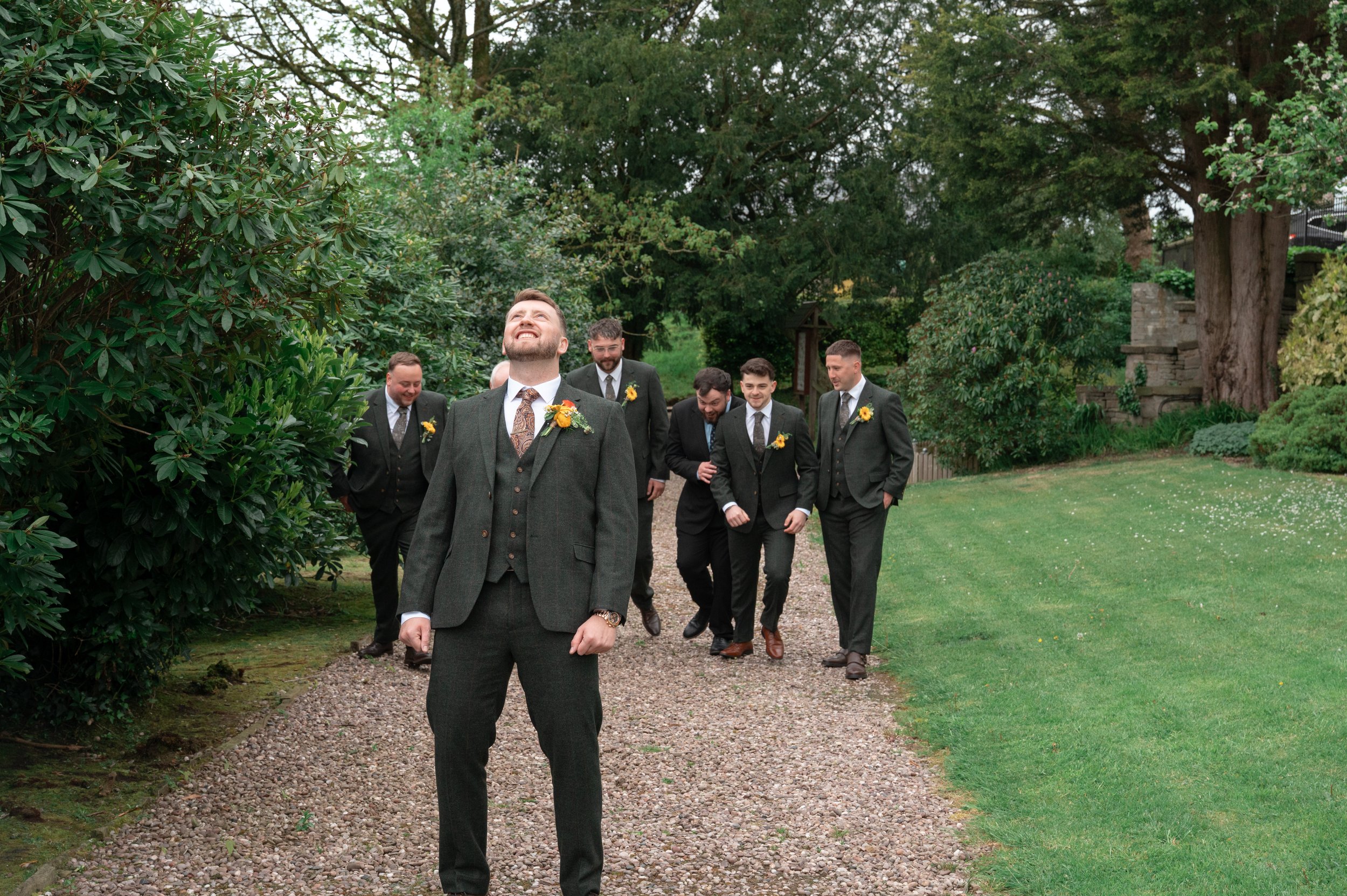 A group of men in suits walking outdoors on a gravel path surrounded by green trees and bushes, with one man in front smiling and looking up.