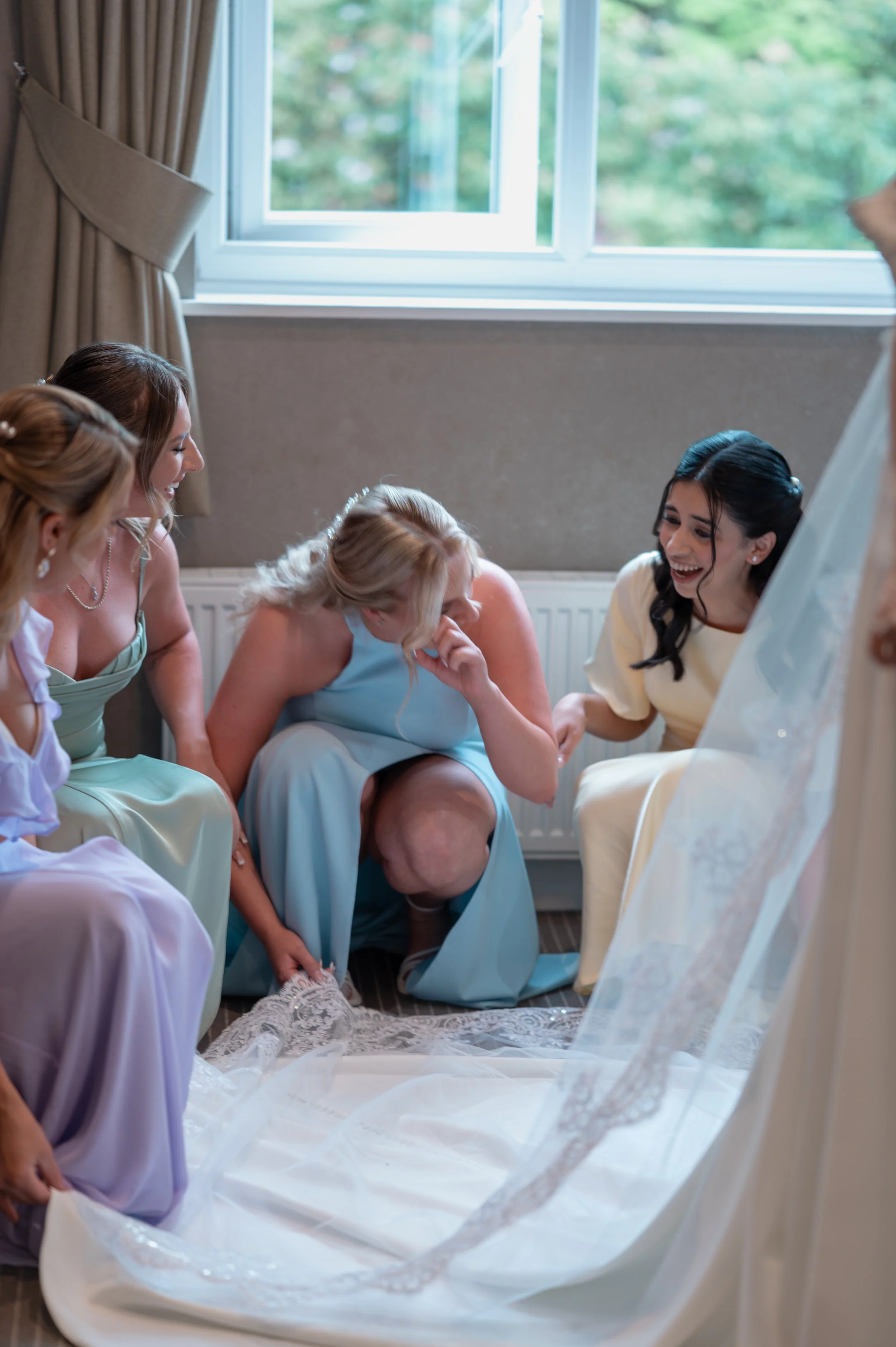 Women in pastel dresses sitting on the floor, laughing and celebrating around a wedding dress laid out on a white cloth, with a large window and beige curtains in the background.