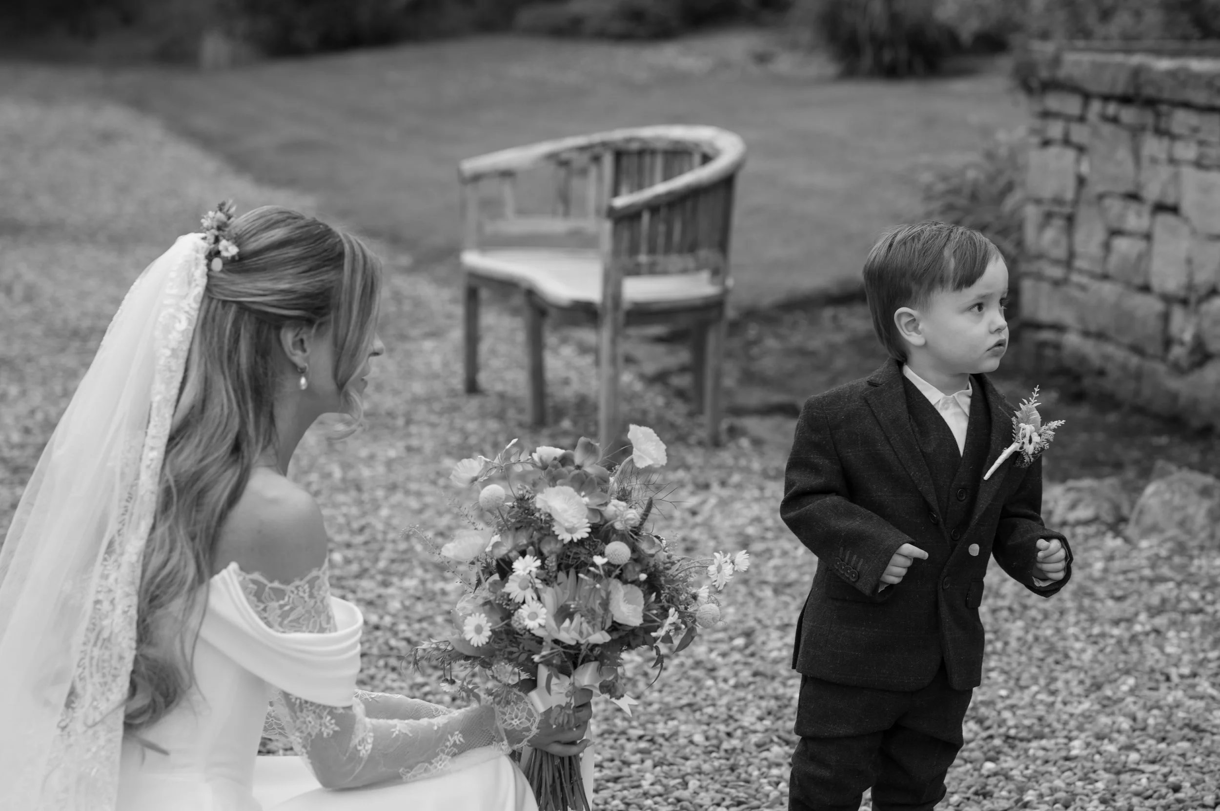 A black and white photo of a woman dressed as a bride holding a bouquet of flowers, and a young boy in a suit holding a small flower, likely at a wedding or special event outdoors.