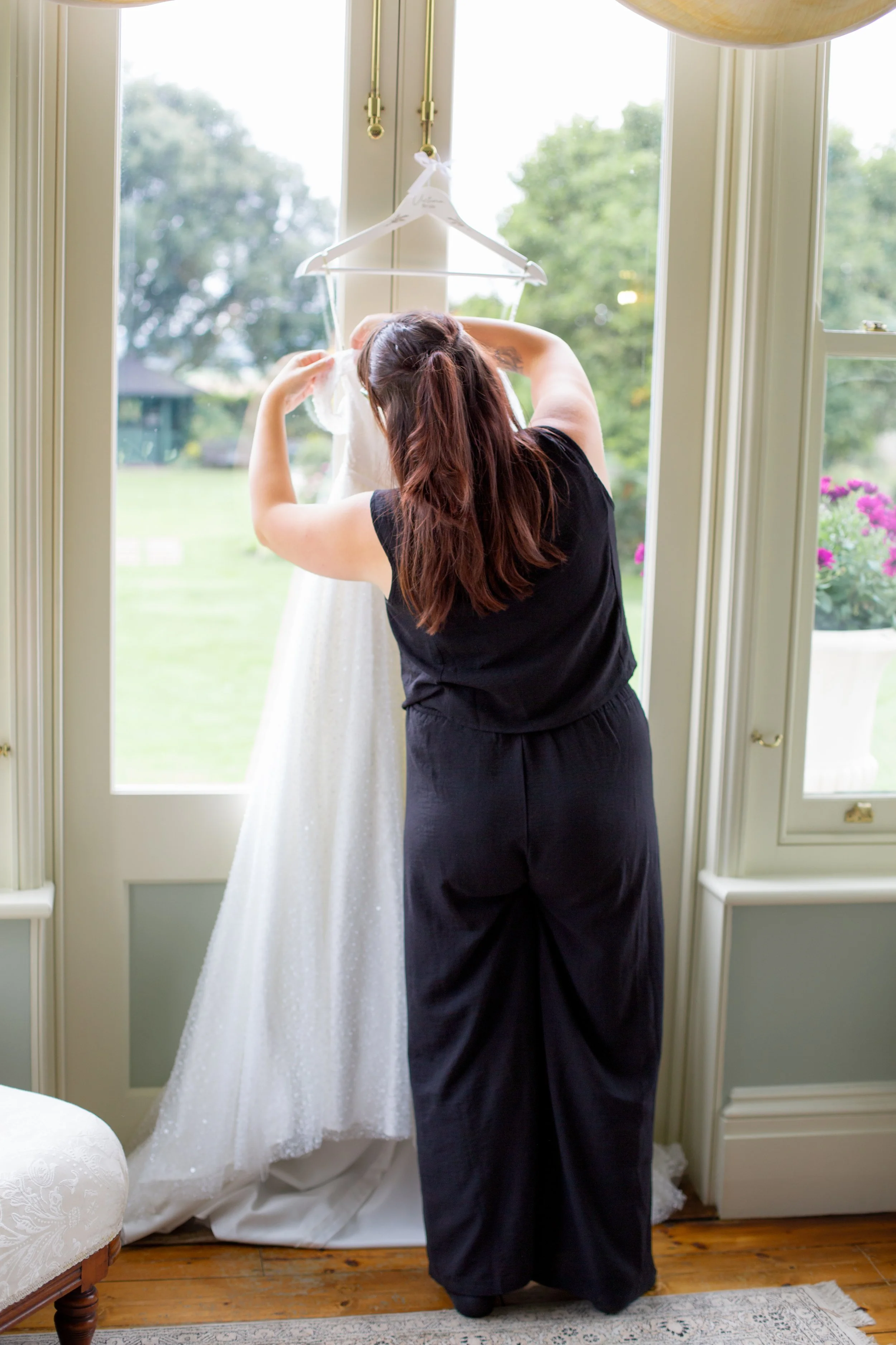 A woman with long red hair dressed in black pants and top is standing inside a room, looking at a wedding dress hanging on a hanger near a window with green trees outside.