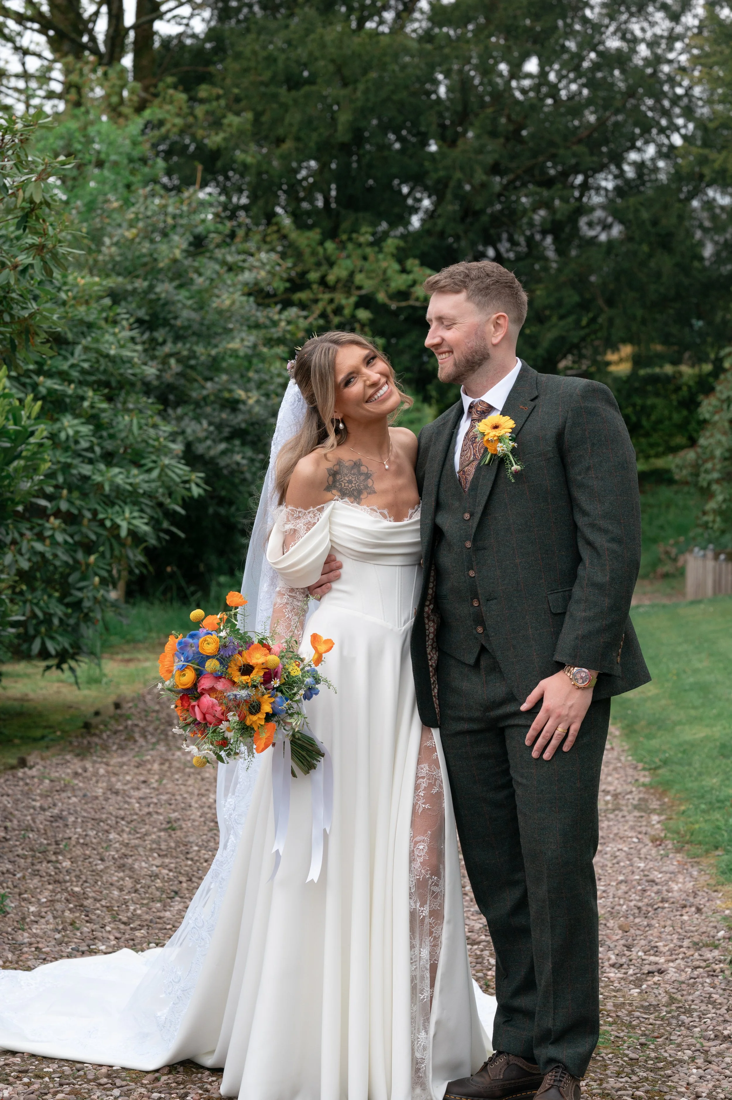 A bride and groom standing outside on a gravel path surrounded by green trees, smiling and looking at each other. The bride is wearing a white wedding gown with lace details and holding a colorful bouquet, and the groom is dressed in a dark plaid suit with a boutonniere featuring a yellow flower.
