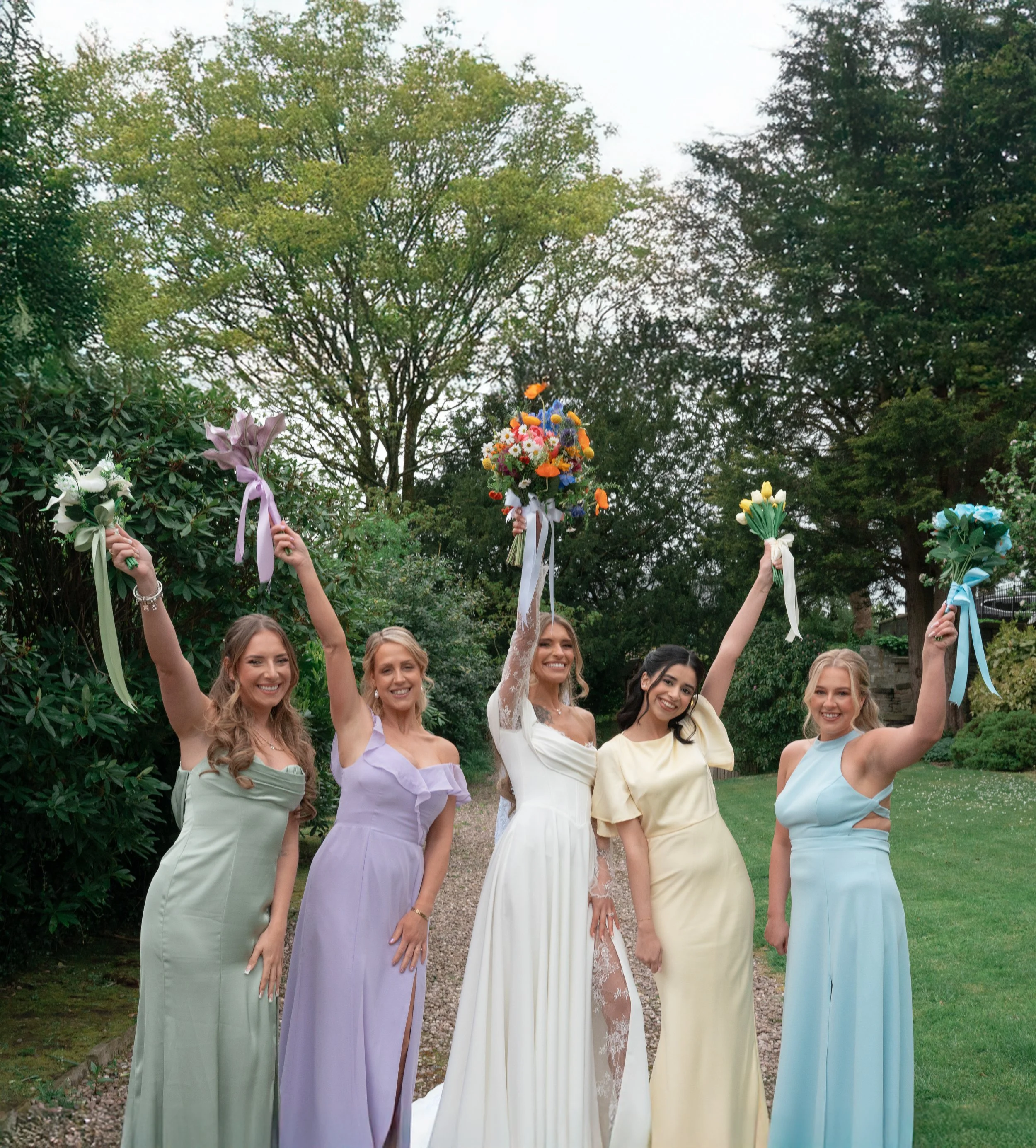 A group of women in pastel-colored dresses holding floral bouquets celebrating outdoors in a garden.