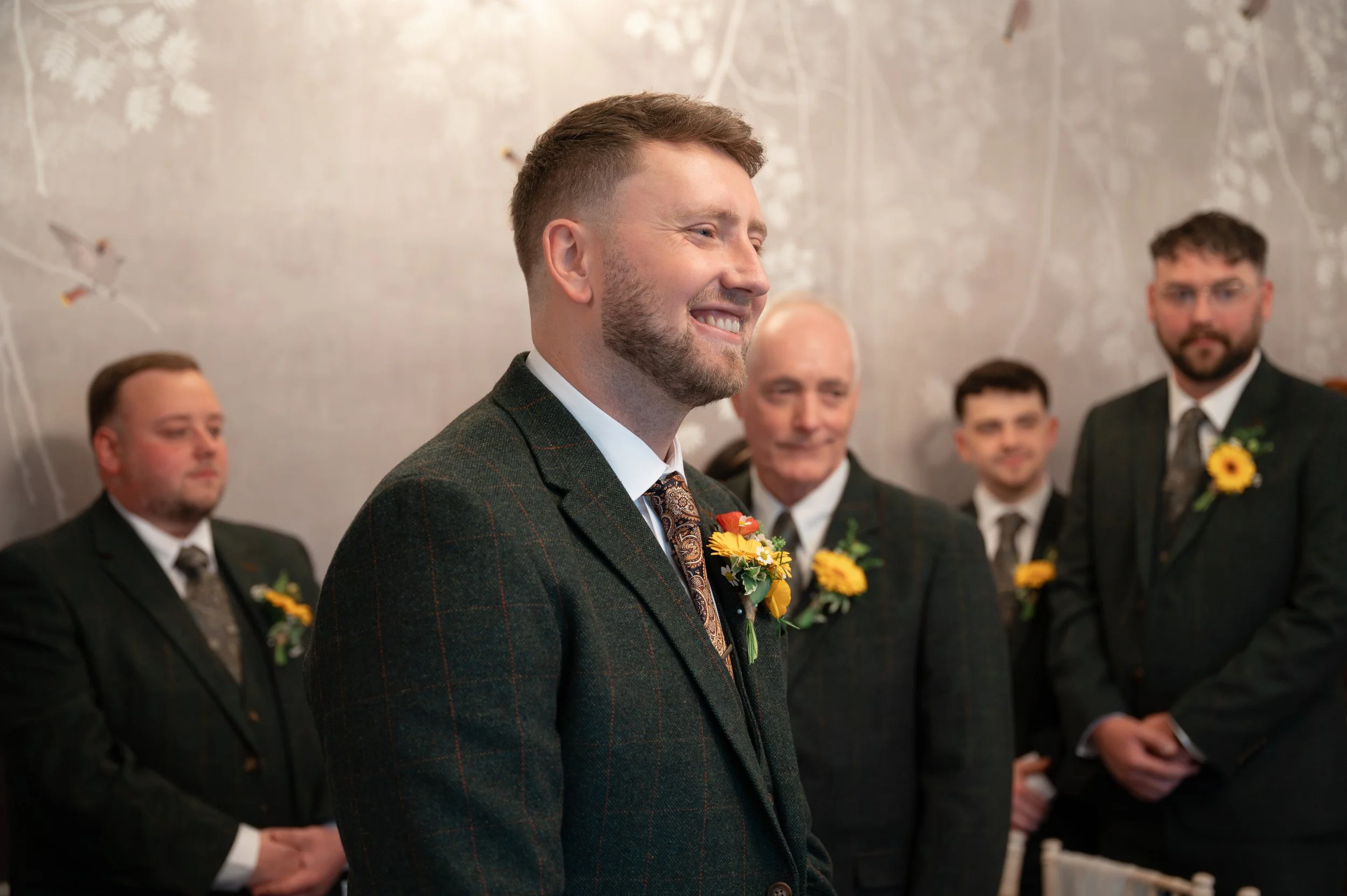 A man wearing a dark green plaid suit and patterned tie, smiling during a wedding ceremony, with a boutonniere of yellow flowers and greenery on his lapel. Other men in suits with similar boutonnieres stand behind him.