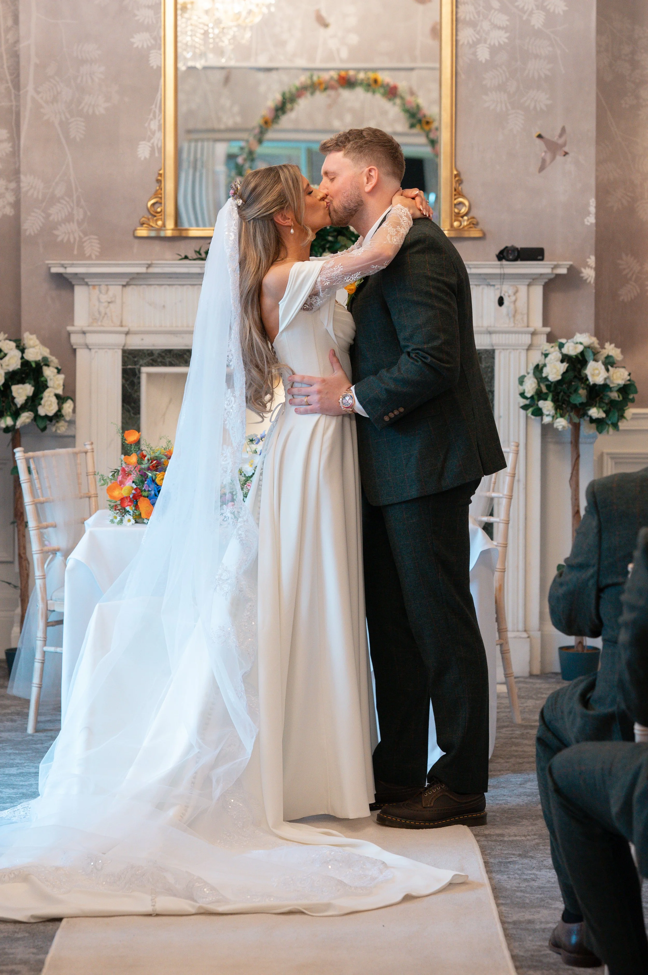 A bride and groom kiss during their wedding ceremony indoors. The bride wears a white gown with lace sleeves and a veil, and the groom wears a dark plaid suit. They embrace in front of a white fireplace with floral arrangements and a large mirror ref