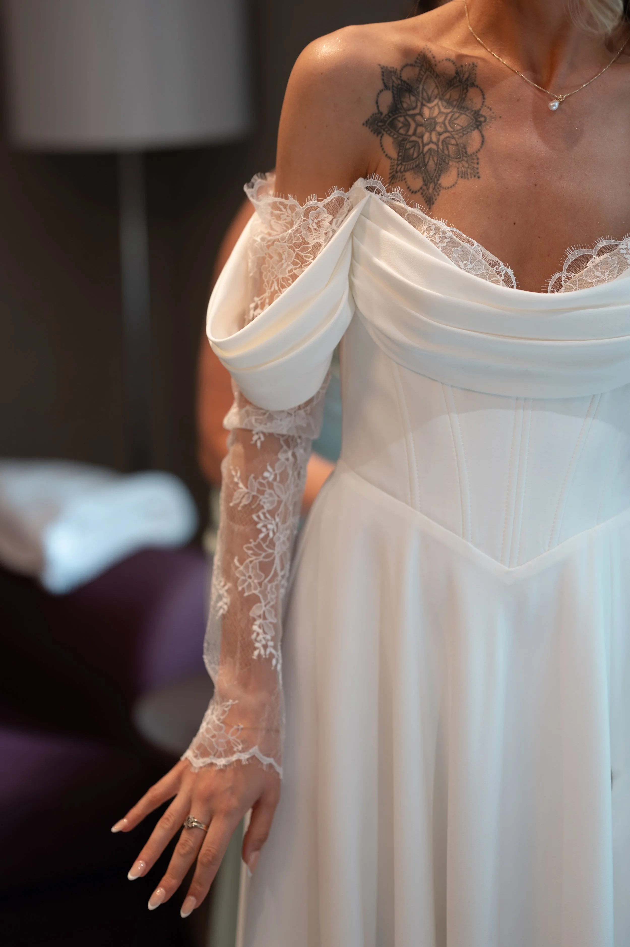 Close-up of a bride’s upper body showing a tattoo on her shoulder, wearing a white wedding gown with lace off-the-shoulder sleeves and a pearl necklace. Her hand with manicured nails is visible.