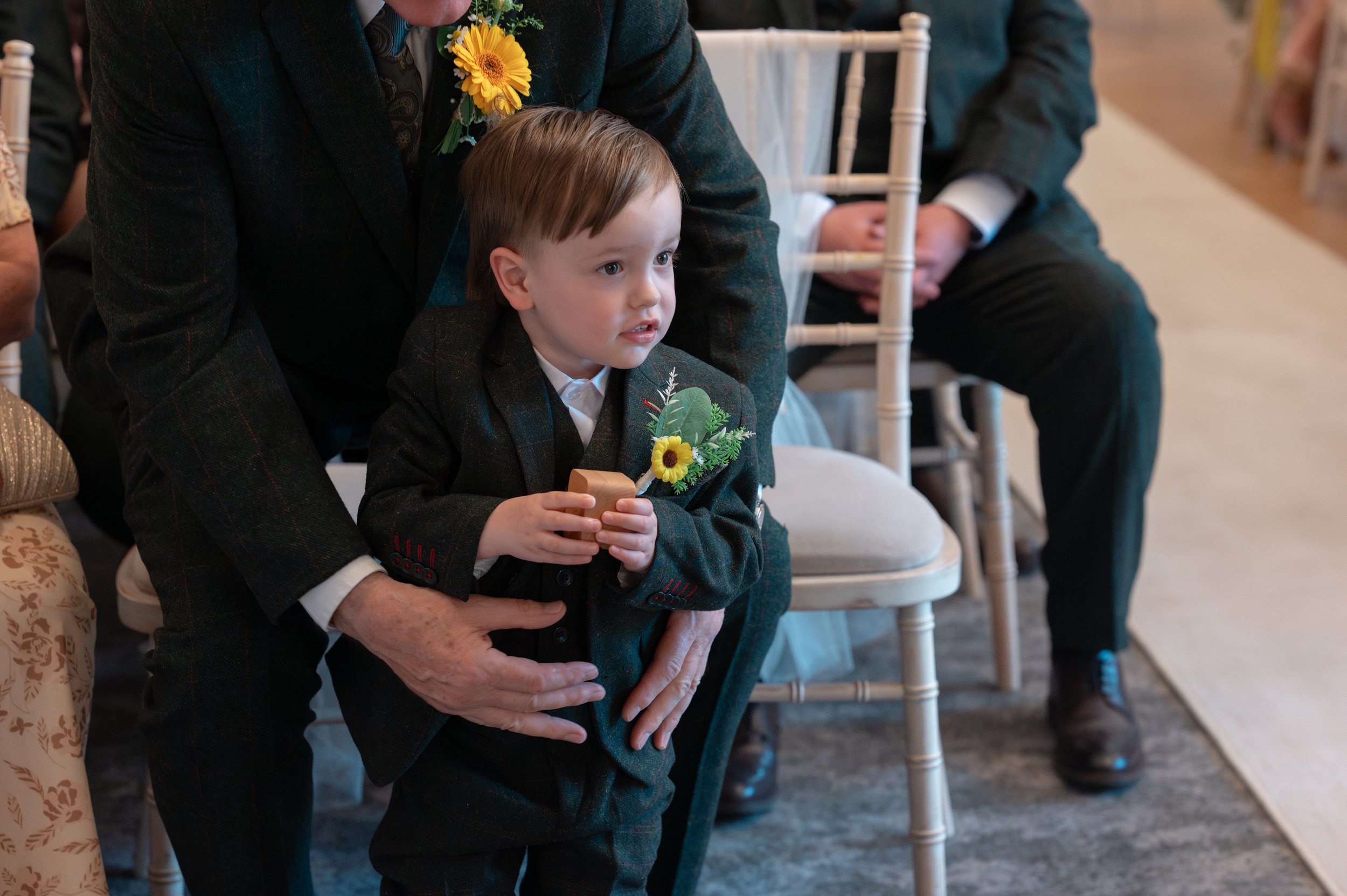Young boy in formal attire with a flower boutonniere, sitting on a white chair at a formal event, surrounded by adults, with one adult's hand on his lap.