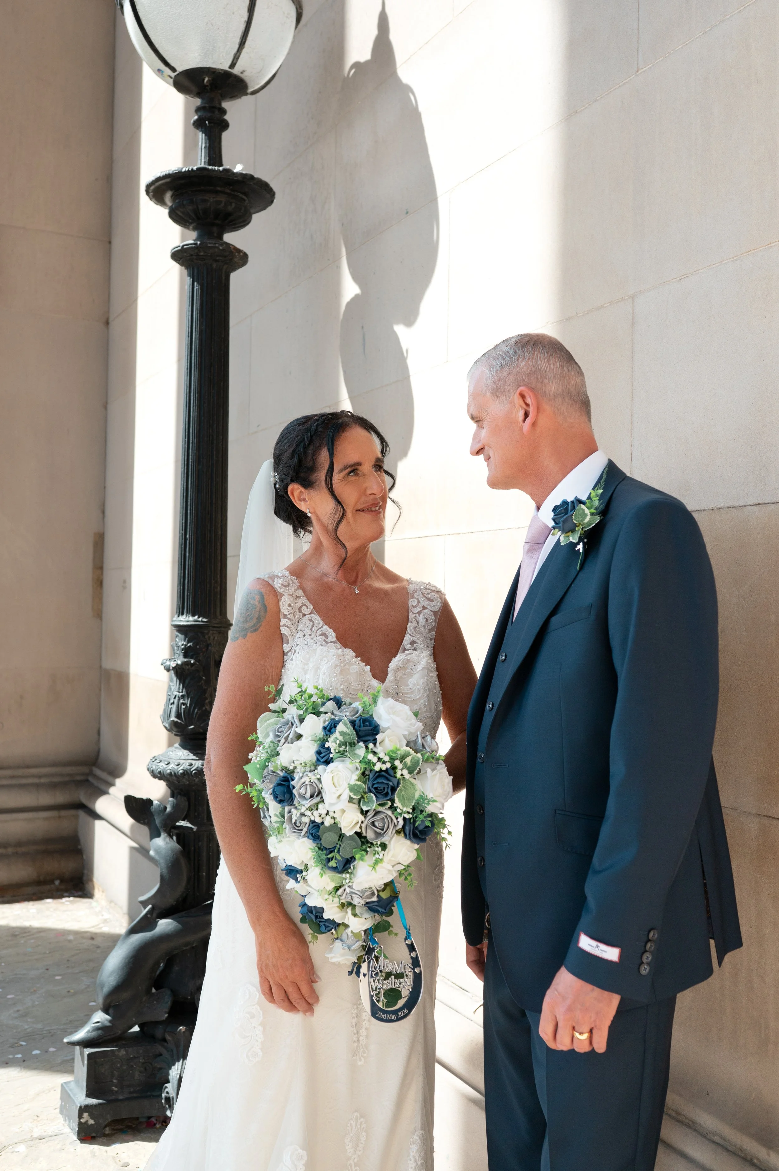 A bride and groom standing in front of a beige wall, looking at each other. The bride wears a white wedding dress and holds a bouquet of white and blue flowers. The groom wears a navy blue suit with a boutonniere. They are outdoors near a black lamppost.